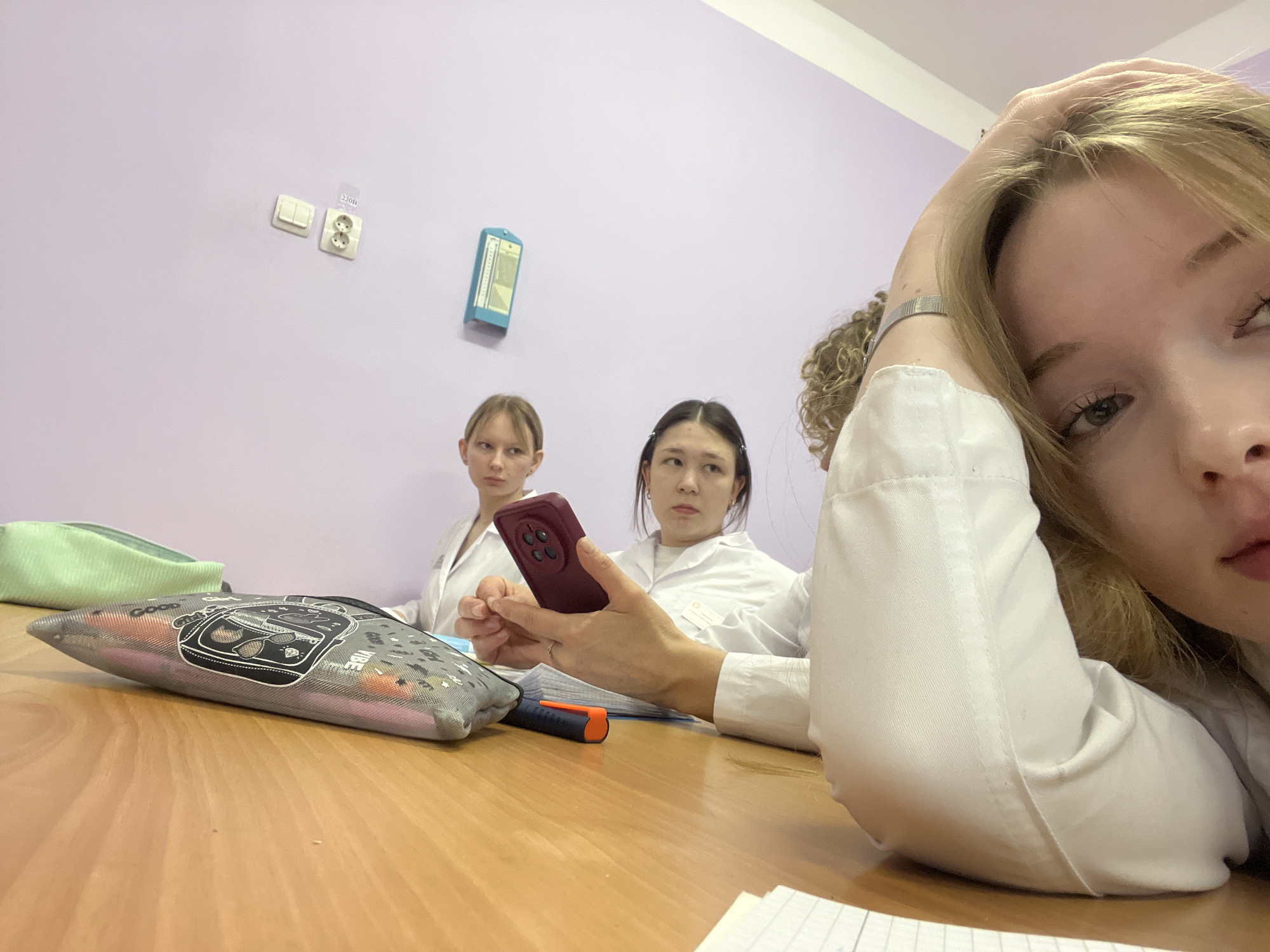 The image depicts a group of three young women sitting at a desk. All appear to be wearing white lab coats. One woman is holding a maroon-colored smartphone. Another woman is looking at the phone. The third woman is positioned closest to the camera, with her head resting on her hand. On the desk, there is a gray pencil case with a black design and a green pencil case. There is also an orange and black marker. The wall behind the women is light purple and has a white light switch and a white electrical outlet on it. A blue wall-mounted chart can be seen hanging on the wall. The setting is the city of Chaikovsky, Russia.