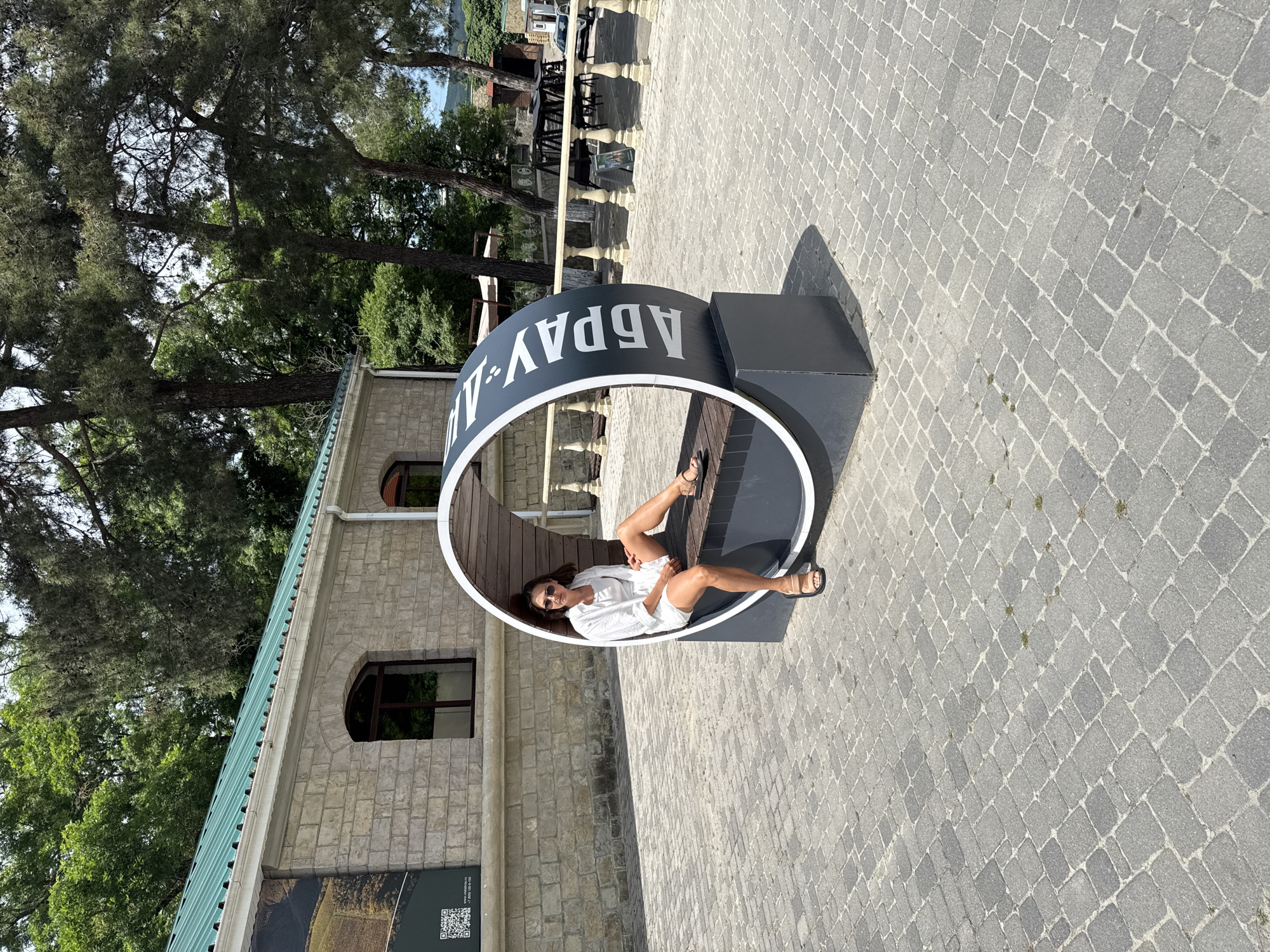 A woman wearing a white shirt and shorts sits on a circular bench with "АБРАУ" written on it in white letters. The bench is made of dark gray metal with wooden seating. She is wearing sunglasses and sandals. The bench is located on a paved area with gray cobblestones. In the background, there is a building with a light-colored stone facade and arched windows. Trees with green foliage are visible above the building and around the area. The scene appears to be in a park or recreational area in Abrau-Dyurso, Russia.