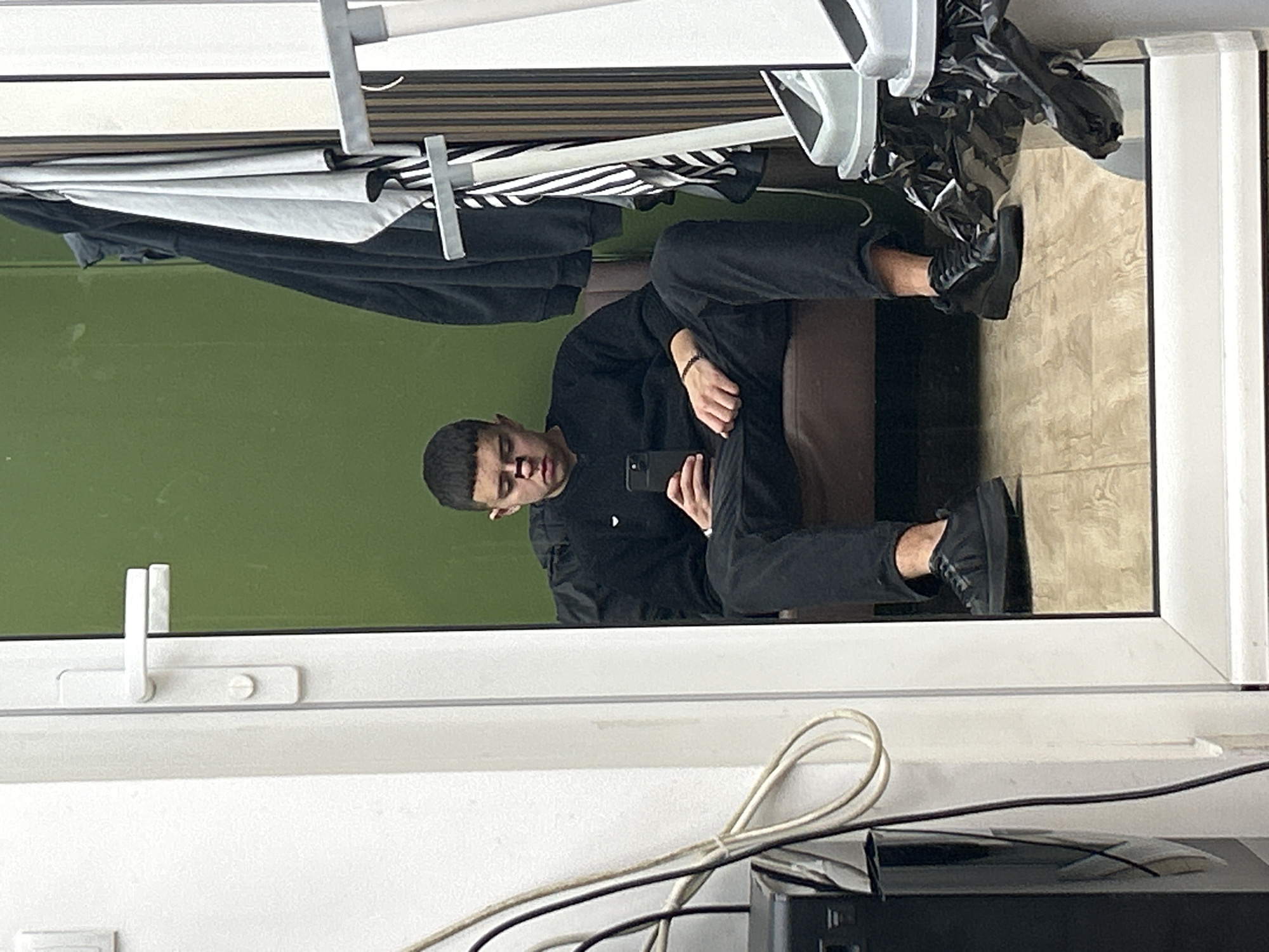 A mirrored image shows a man sitting on a brown chair, wearing a black shirt, black pants, and black shoes, and holding a phone. The reflection is in a window, with a green exterior visible behind the window. Above the man in the reflection, clothes hang on a rack. Below the window in the foreground are white window frames and electrical cords connecting to a black object. The reflection indicates the photo was taken inside an indoor space, and there is a white tiled floor. There is no information in the image to verify its location in Zlatograd, Bulgaria.