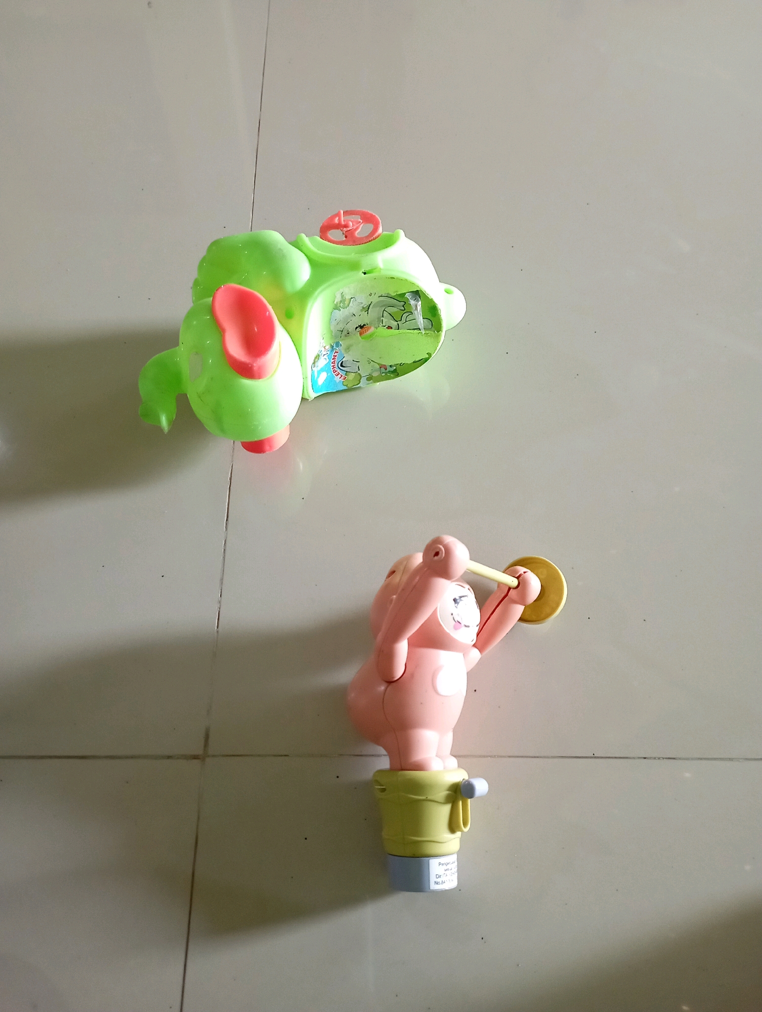 Two toys are on a tiled floor. In the upper part of the image, a green toy elephant with red ears and feet is lying on its side. The elephant has a clear plastic section on its back that contains a colorful scene. A red steering wheel is visible on top of the elephant's head. In the lower part of the image, a pink toy figure, resembling a pig, stands upright in a yellow bucket. The figure is holding a yellow cymbal in its right hand and appears to be about to strike it with a stick. The floor is tiled in a neutral color and shows grout lines. The lighting suggests an indoor environment, possibly a home. There is no indication of the city of Sonobekel, Indonesia, in the image.