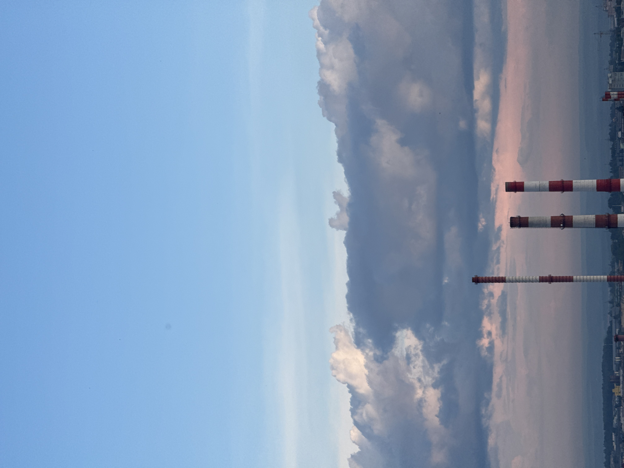 The image is a vertically oriented photograph depicting a wide sky filled with clouds and a landscape below. The sky on the left is a clear, bright blue, transitioning to a more muted, overcast sky on the right, filled with dark, heavy clouds tinged with pink and orange hues, suggesting either sunrise or sunset. In the middle ground, several industrial smokestacks, painted with red and white stripes, rise above a distant cityscape. These smokestacks are a prominent feature, indicating an industrial or power generation area. Below the smokestacks, a blurred landscape of buildings and trees can be seen. The overall mood is somewhat dramatic due to the contrast between the clear blue sky and the ominous clouds, and the presence of the industrial structures. The time of day is likely either dawn or dusk. There is no visible text in the image.