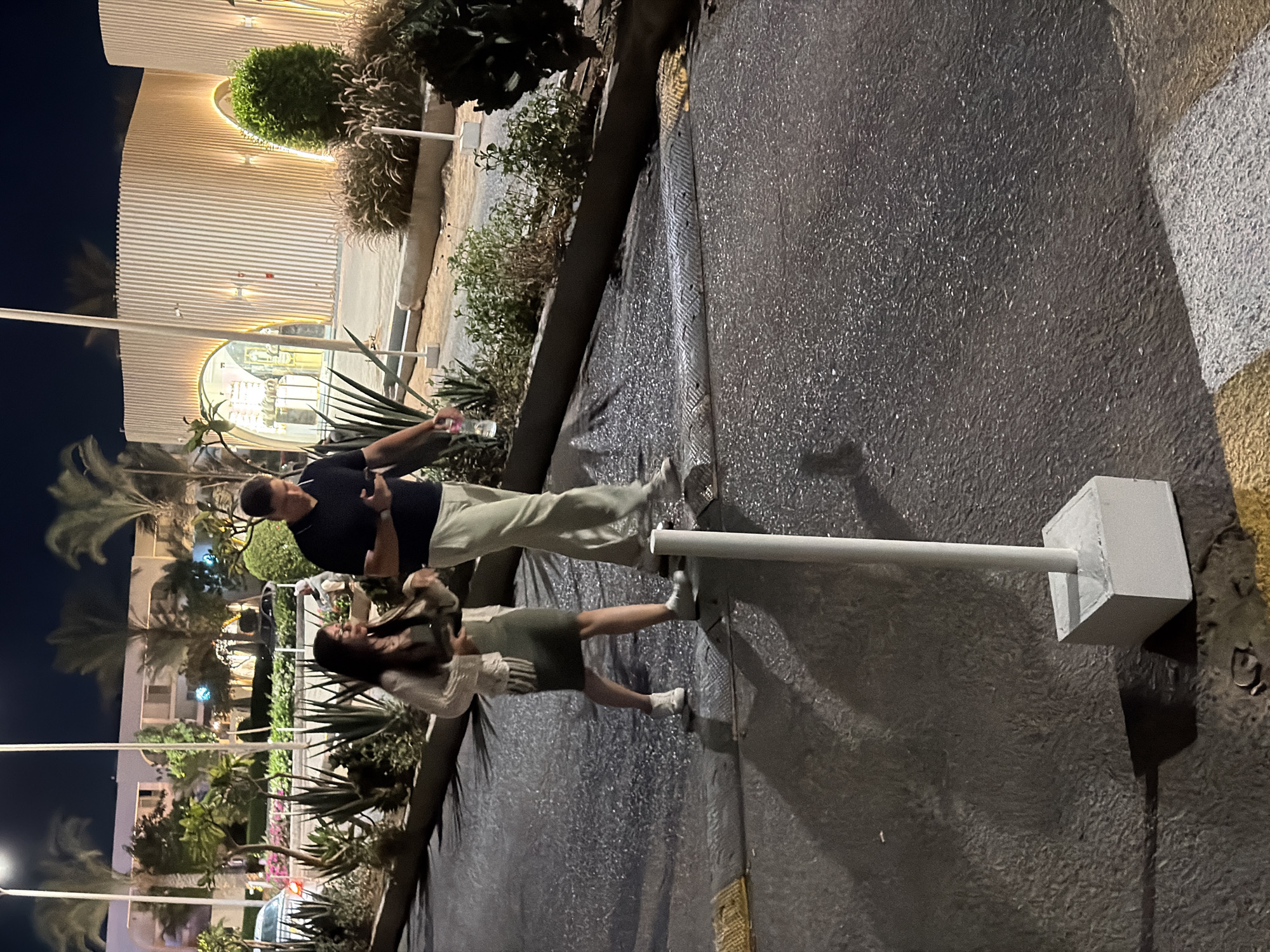 An outdoor night scene in Sharm El Sheikh, Egypt, shows a man and a woman walking on a paved surface. The man, wearing a dark polo shirt and light-colored pants, holds a smartphone in his right hand and a water bottle in his left. The woman, dressed in a light-colored cardigan over a green dress, carries a clutch bag. They are walking side-by-side. In the background, palm trees and modern architecture with illuminated vertical slats suggest a resort or commercial area. The ground is wet, reflecting the ambient light. A white pole on a concrete base stands in the foreground, casting a shadow on the wet pavement. Yellow and white markings are visible on the road surface.