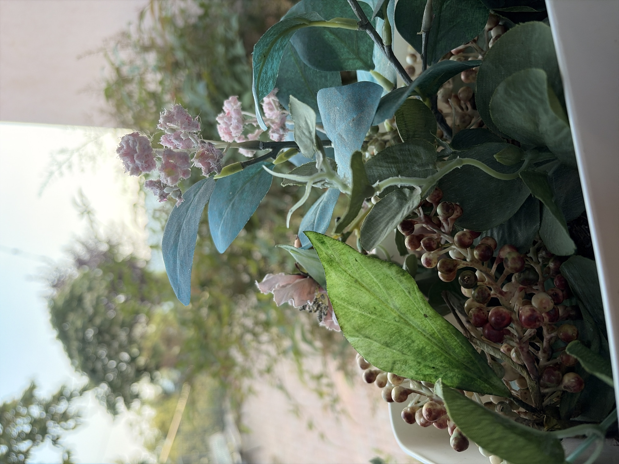 This is a close-up, eye-level, vertical shot of an artificial floral arrangement placed near a window. The arrangement features a variety of elements, including large, muted blue-green leaves, smaller pinkish-purple clusters of flowers, and branches dotted with small, bead-like berries in shades of cream, burgundy, and green. A prominent, textured green leaf with visible veins is positioned in the foreground, slightly to the right of center.
The background is softly blurred, suggesting an outdoor scene viewed through the window. Hints of trees and foliage, bathed in bright, diffused light, indicate it is daytime, likely sunny, with clear skies visible. The soft focus and the interplay of light and shadow on the artificial flowers and leaves create a gentle, somewhat serene atmosphere. The arrangement appears to be indoors, possibly in a home or a commercial space in Cornedo Vicentino, Italy, given the provided location context. There is no visible text in the image.
