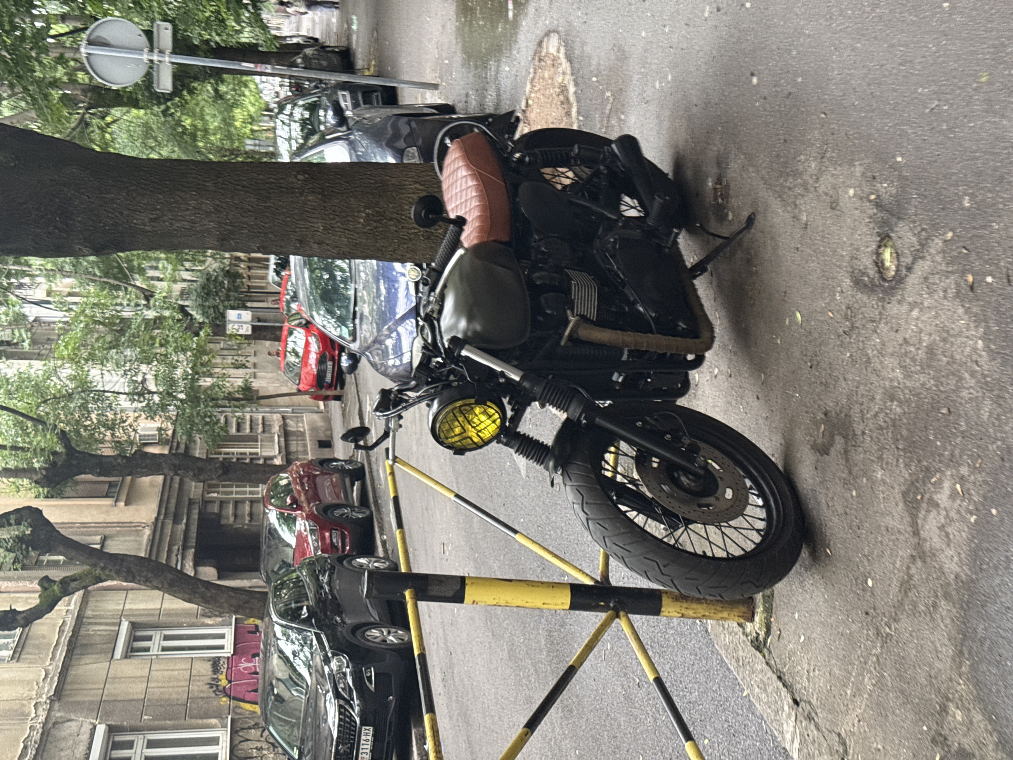 The image depicts a street scene in Belgrade, Serbia, showcasing a parked motorcycle adjacent to a black and yellow barricade. The motorcycle features a tan leather seat and a yellow-tinted headlight. Several cars are parked along the street, with the visible license plate "XH 3116-HX" on a black SUV. A tree trunk is prominently positioned beside the motorcycle and cars. The background includes buildings with visible windows and tree foliage. A street sign is mounted above the tree. The setting appears to be an urban environment during daylight.