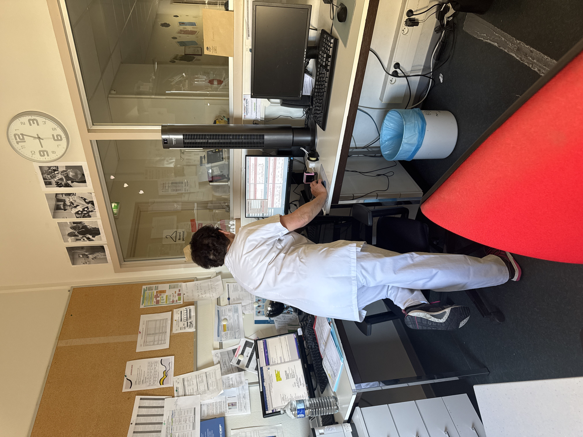 A woman in a white medical uniform, facing away from the camera, stands behind a desk with two computer monitors. She is positioned between the two monitors, with her left hand resting on the keyboard of the left computer. She is wearing dark athletic shoes. The desk is equipped with a keyboard, mouse, and a water bottle. A tall, black tower fan stands to the right of the left monitor. The office environment features a corkboard covered in papers on the left wall, a clock on the wall above the desk, and a glass partition separating this area from another room. A blue trash can is situated on the floor to the right of the desk. The scene appears to be indoors, likely in a medical or administrative office setting. No specific location in Tourcoing, France is discernible from the image itself.