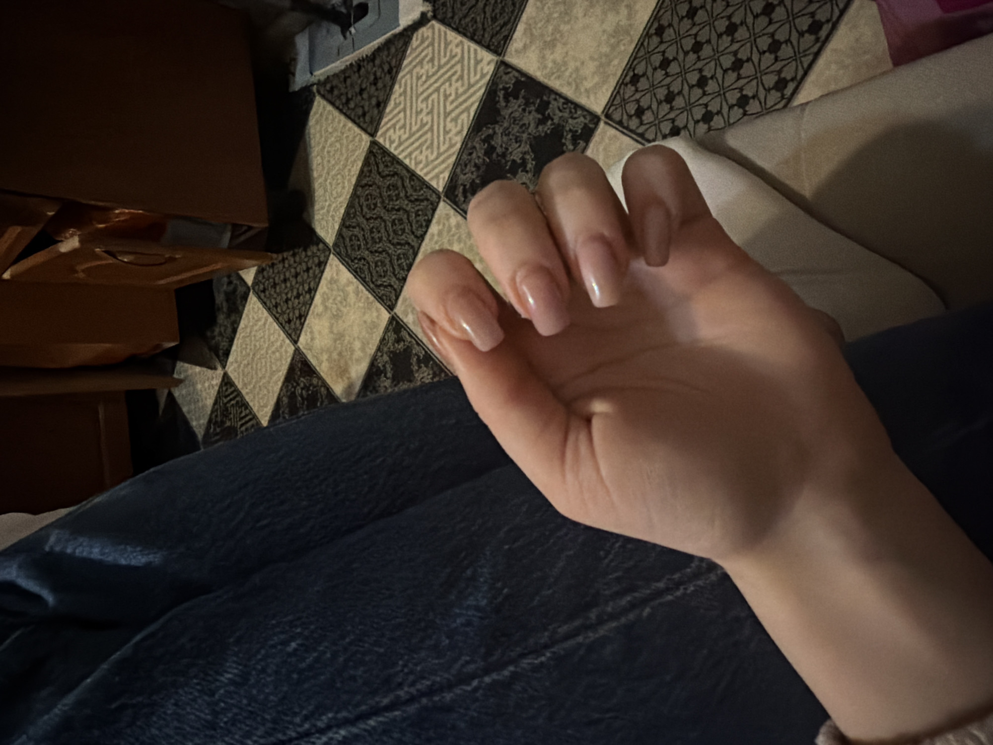 A close-up shot focuses on a person's left hand, adorned with light pink, almond-shaped acrylic nails, resting against a thigh covered in dark blue denim. The background reveals a black and beige patterned tile floor, with a wooden cabinet or furniture piece partially visible to the right. The lighting is dim, casting soft shadows across the hand and the floor. The scene appears to be indoors, likely in a residential setting.