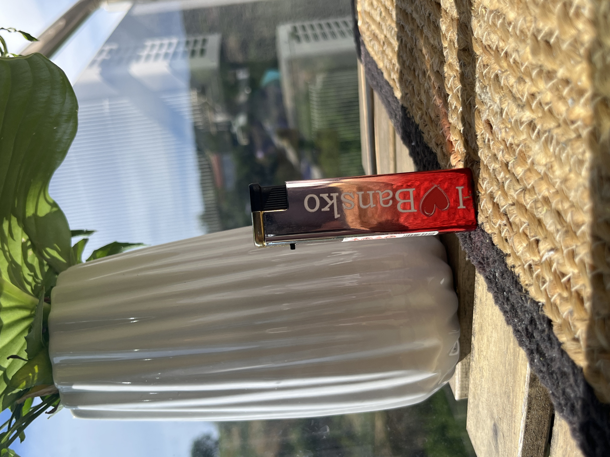 A metallic red lighter with white text reading "I ❤️ Bansko" lies on a woven, light brown mat. The lighter is positioned vertically, with its top visible and its body extending downwards towards a white ribbed vase. The vase contains a leafy green plant, its leaves casting shadows on the vase's surface. The scene appears to be outdoors, possibly on a balcony or windowsill, with a bright, sunny sky and a blurry background of buildings and trees, suggesting a daytime setting. The overall mood is relaxed and casual.