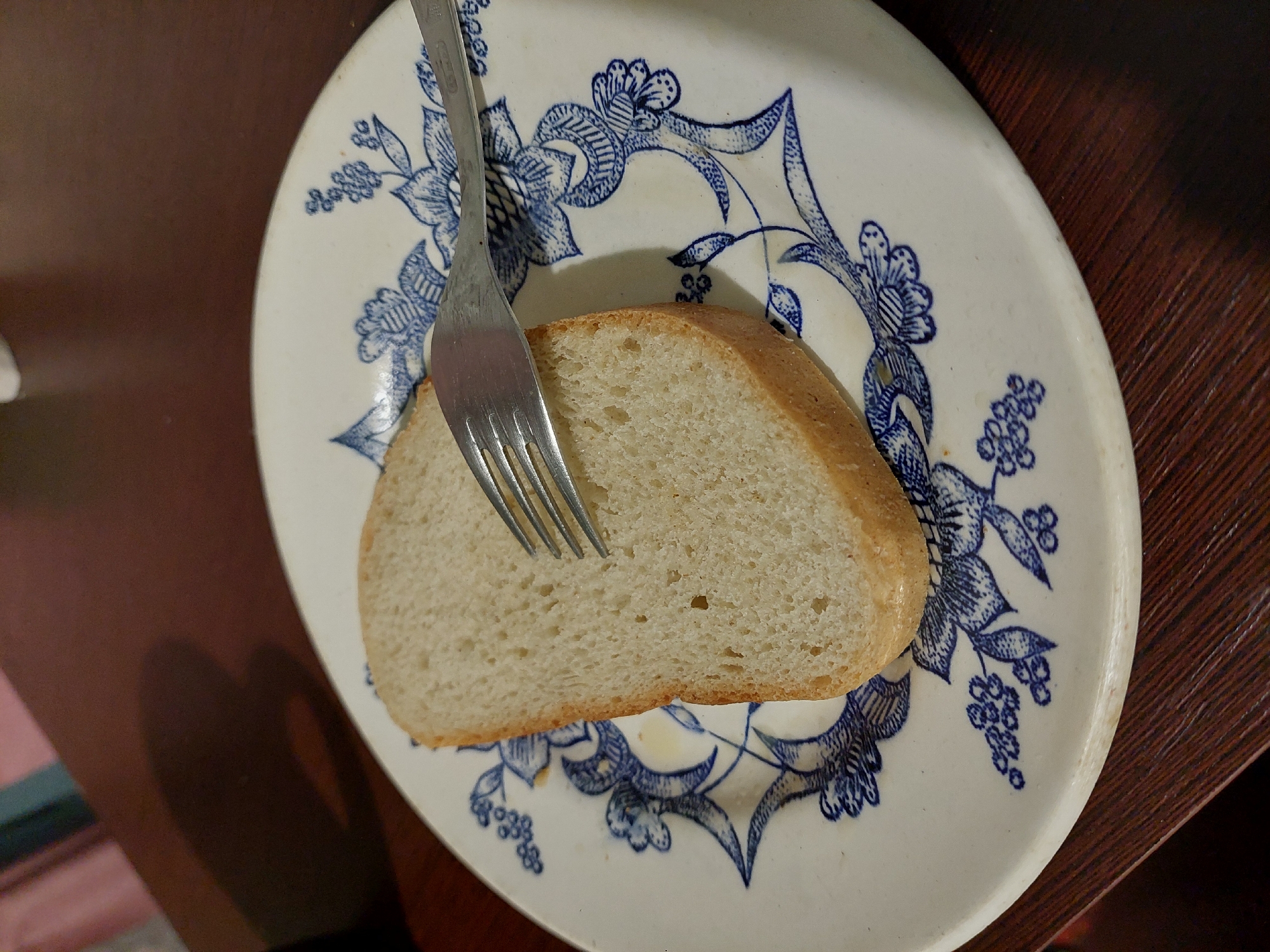 A close-up shot shows a slice of white bread on a white plate with a blue floral pattern. A silver fork is embedded in the bread, with its tines visible. The bread appears to be a simple loaf, with a light brown crust and a soft, airy interior. The plate has a decorative border with intricate blue floral designs, typical of traditional Ukrainian ceramics. The background is dark and out of focus, suggesting the photo was taken indoors, possibly on a wooden table. The lighting appears to be artificial, casting a shadow on the left side of the plate. The image is clear and well-lit, highlighting the texture of the bread and the details of the plate. There is no visible text in the image. The setting suggests a casual meal or snack.