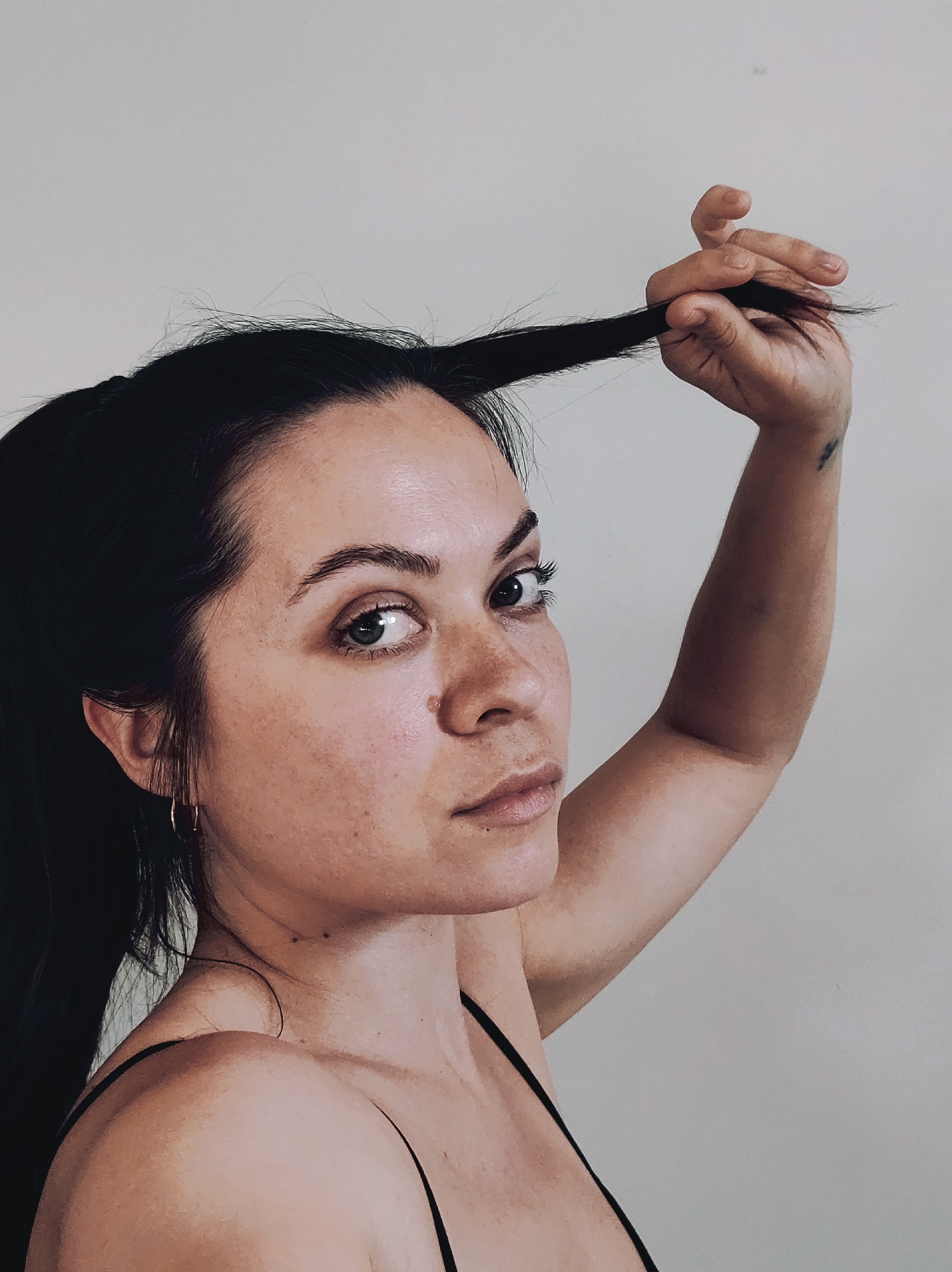 A medium shot captures a woman against a plain, light-colored wall. The woman, with fair skin, dark hair pulled back into a ponytail, and dark eyebrows, is looking directly at the camera. Her left arm is raised, holding a section of her hair with her hand. She wears a simple black spaghetti strap top. A small gold hoop earring is visible on her left ear. A tattoo is visible on the inside of her raised arm. The lighting is even and soft, illuminating her features and the background uniformly. The location is unknown.