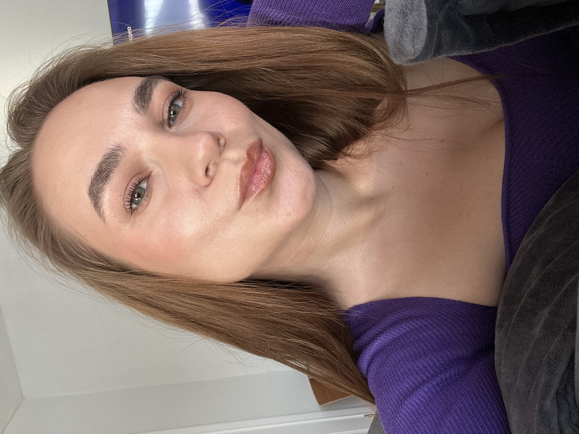 A close-up, angled selfie of a woman. She has light brown hair and green eyes. She is wearing a purple knit shirt. Part of a gray blanket is visible. The background is a white wall. The location is not discernible from the image.