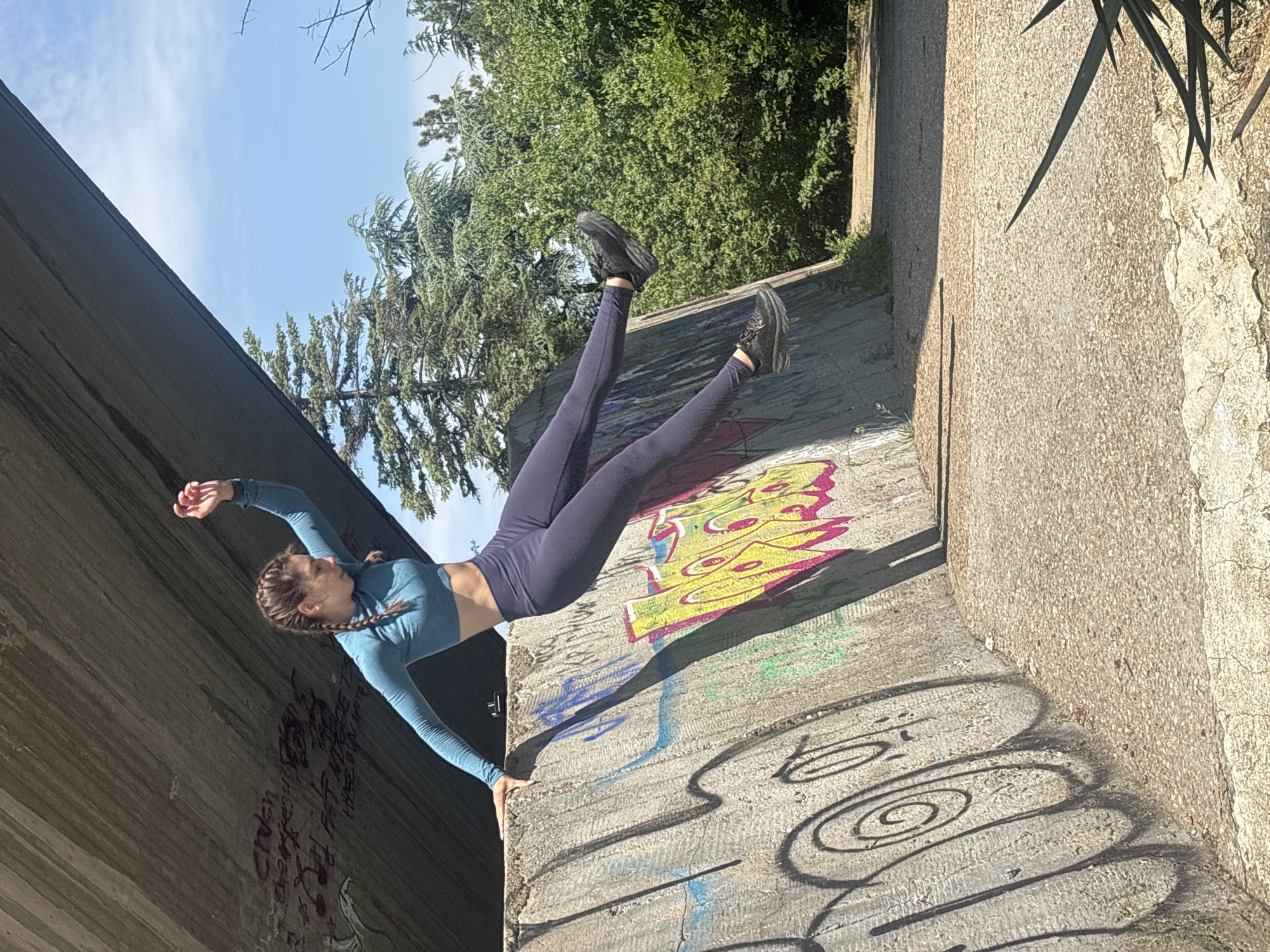 An outdoor scene captures a young Caucasian female performing a dynamic maneuver under a concrete bridge or overpass, characteristic of the area near the Asparuhov Bridge in Varna, Bulgaria. The subject, with dark braided hair, wears a teal long-sleeved crop top and dark blue leggings, along with black athletic shoes. She is depicted in a horizontal, suspended posture, extending her legs to the right while her left hand is placed on a graffiti-covered, slanted concrete wall. Her right arm is raised and bent.
The environment consists of rough, textured concrete structures. A large, grey underside of the bridge occupies the upper left, bearing red and black graffiti, including script and musical note motifs. Below this, a heavily graffitied concrete retaining wall, featuring yellow, pink, blue, green, and black spray paint in various styles (e.g., "bubble letters" and spirals), slopes downwards to the right. The ground is a light-colored, pebbled concrete path showing cracks. Beyond the structures, a bright blue sky with sparse white clouds is visible, along with dense green vegetation, including coniferous trees and bushes, suggesting a park or natural area. Strong sunlight casts prominent shadows from the structures and the individual, indicating a clear day.