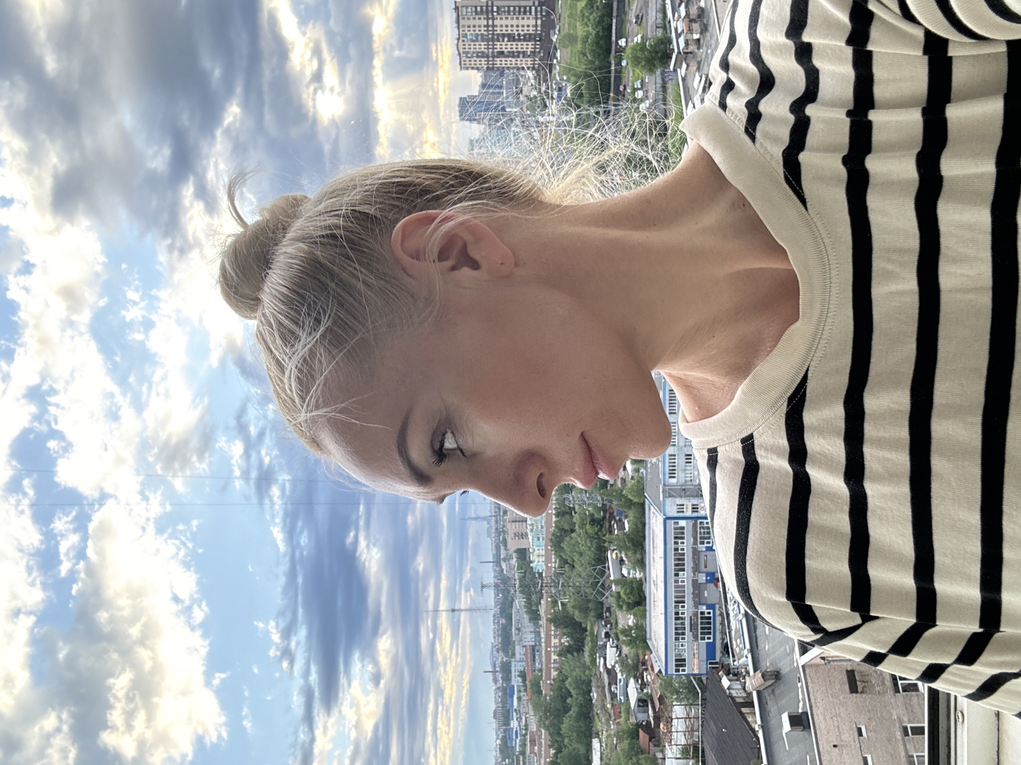 A woman with blonde hair in a bun is shown from the chest up in a profile view, looking to the left. She is wearing a black and white striped t-shirt. Behind her, a cityscape with buildings, trees, and power lines is visible under a cloudy sky with rays of sunlight breaking through. The image appears to be taken from a high vantage point.