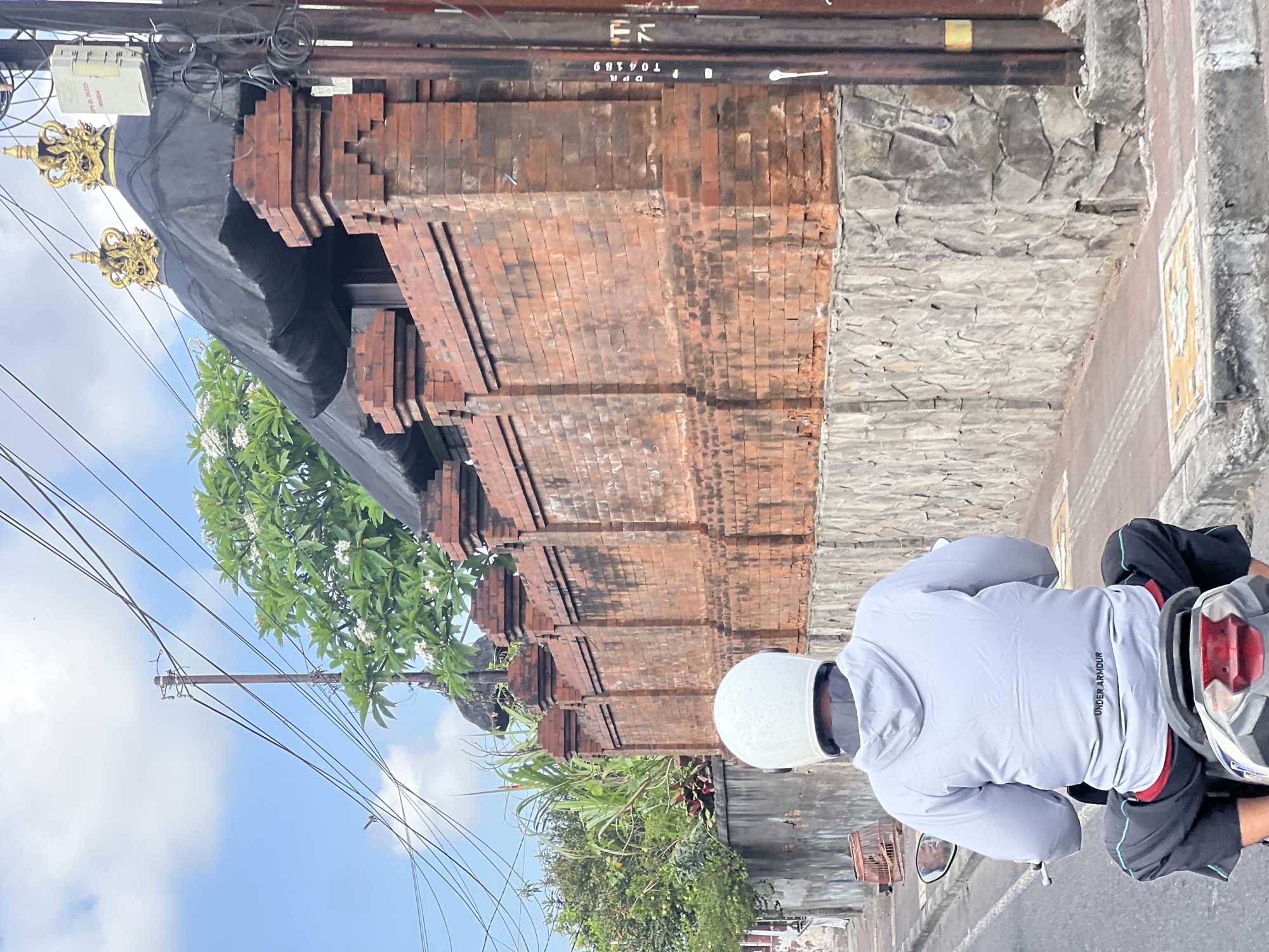 The image depicts a person riding a scooter on a street in Denpasar, Indonesia. The rider is wearing a white helmet and a light gray hoodie with "UNDER ARMOUR" visible on the back. They are also wearing black shorts with teal trim and a red undershirt. The scooter has a red taillight and a license plate that is partially visible.
The street is paved and has a sidewalk with decorative tiles. On the right side of the street is a tall brick wall topped with a dark, thatched roof structure adorned with two golden crowns. The wall has a section of rough-hewn stone at its base. To the left of the wall, there are trees and vegetation, including a birdcage on the side of the road. The sky is blue with scattered clouds, and overhead power lines are visible. A utility pole with wires and an electrical box is also present on the right side of the frame. The overall scene suggests a typical street view in a tropical urban environment.