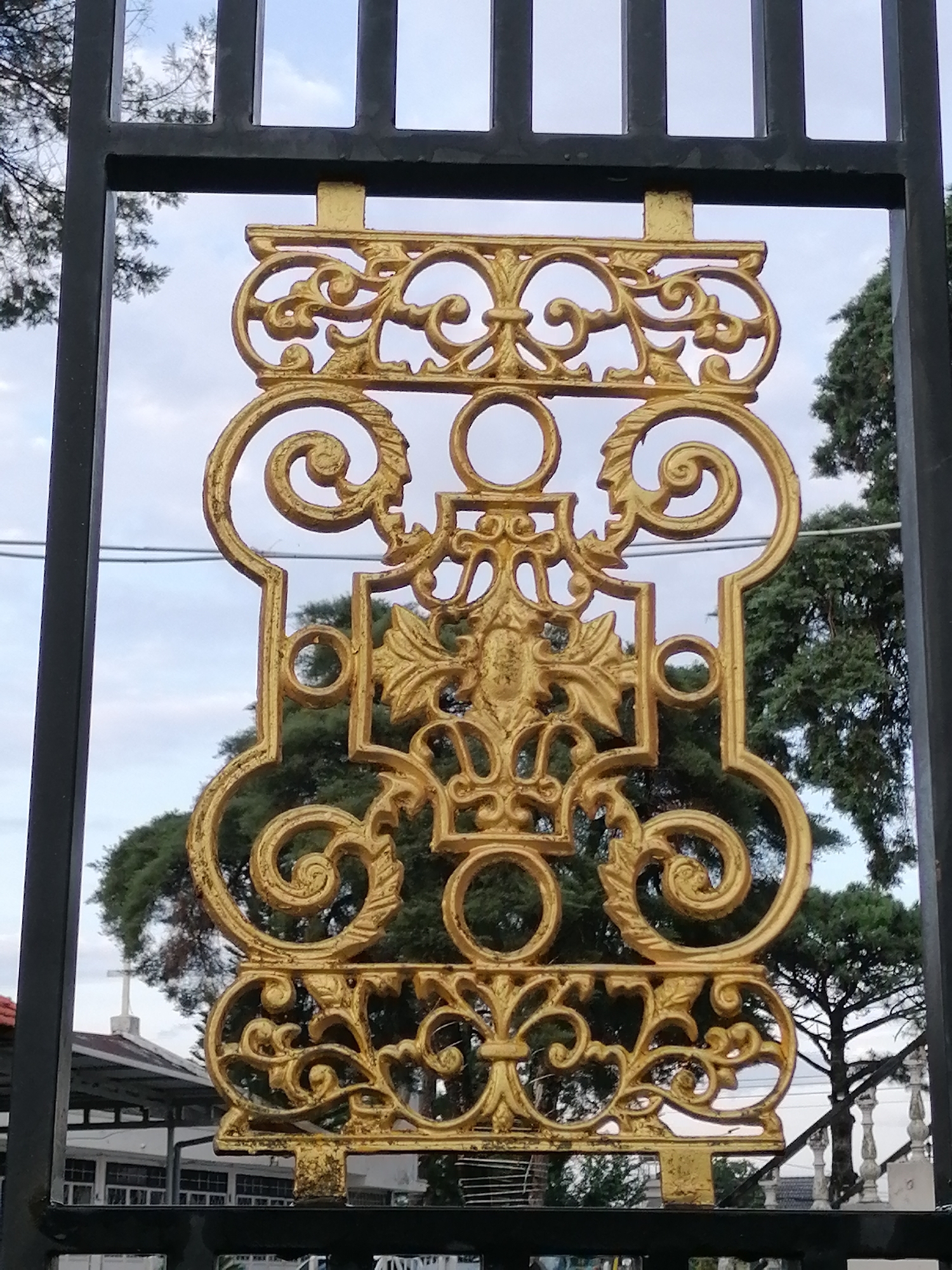 A close-up, head-on shot captures an ornate, gold-colored metal gate in the city of Lâm Viên - Đà Lạt, Vietnam. The gate features intricate scrollwork, symmetrical patterns, and circular elements, set against a backdrop of a cloudy sky and lush green trees. In the distance, a white cross atop a building suggests the presence of a church or religious institution. The black metal bars of the gate frame the elaborate golden design.