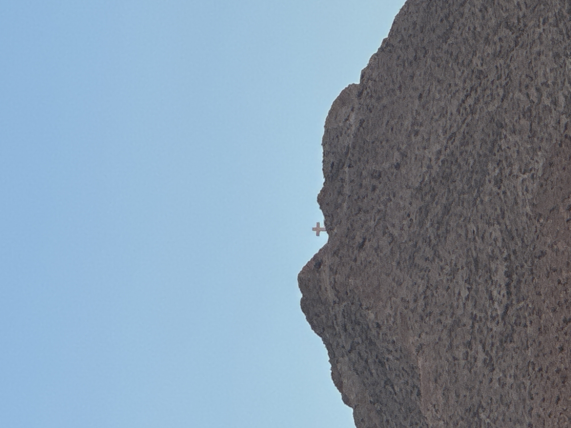 A lone cross stands atop a rocky mountain peak against a clear, pale blue sky. The mountain is covered in dark, rough terrain, with the cross positioned slightly to the right of the center of the frame, its silhouette visible against the sky. The image appears to be a photograph taken from a distance, capturing the vastness of the landscape and the isolated prominence of the cross. The location is identified as Sunland Park, United States of America.