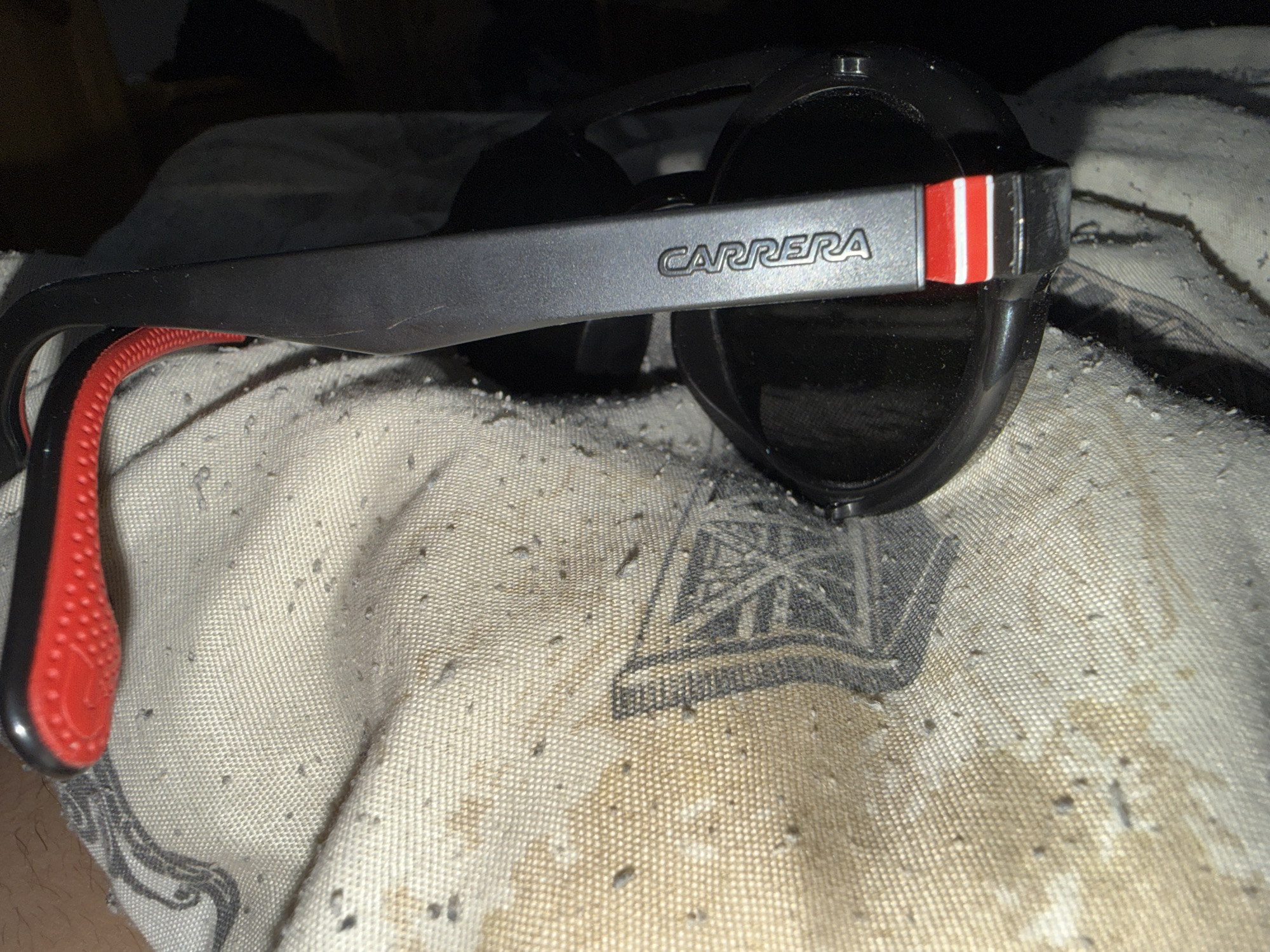 The image shows a pair of black Carrera sunglasses with a gray frame and red accents on an off-white fabric surface with a gray pattern. The sunglasses are positioned with the side bearing the "CARRERA" logo facing upwards. The fabric exhibits worn patches and possible stains. The sunglasses' lens reflects very little light and appear dark. There is no information available to determine the image's location in Burgas, Bulgaria. No people or interactions are shown.