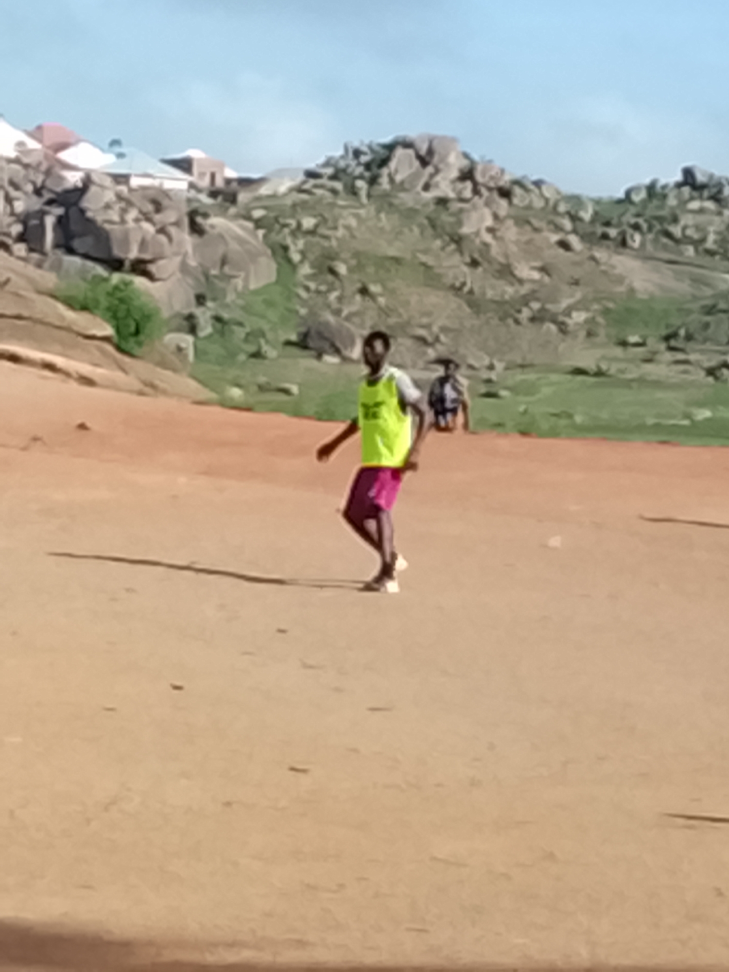 In Jos, Nigeria, a young Black man wearing a bright yellow vest and dark shorts is playing soccer on a dusty, reddish-brown field. The sky is a clear, pale blue, and the background is a rocky, arid landscape with scattered green vegetation and a few houses with light-colored roofs. Another man, wearing a light-colored shirt, is visible in the background, also on the field. The man in the yellow vest appears to be mid-stride, perhaps about to kick a ball, with his body angled slightly to the left. His face is turned to the right, and his expression is not clearly discernible. The scene suggests a casual game of soccer in an open, undeveloped area.