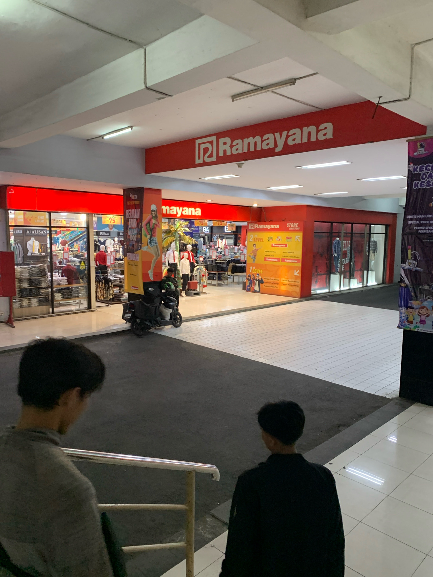 The image shows an interior view of a Ramayana department store, likely located in Malang, Indonesia, as suggested by the signage. Two young males are seen from behind, standing on a dark grey paved area in the foreground, separated by a metal railing. The store itself is visible in the background, featuring bright red exterior walls and large glass windows displaying clothing. A prominent red sign with "Ramayana" in white lettering is mounted overhead. Inside, mannequins and racks of clothing are visible, along with promotional posters and store directories. A black scooter with a rider wearing a helmet is parked to the left of the store entrance. The flooring consists of both dark grey pavement and white tiled areas. Lighting appears to be a combination of overhead fluorescent lights and illuminated signs.