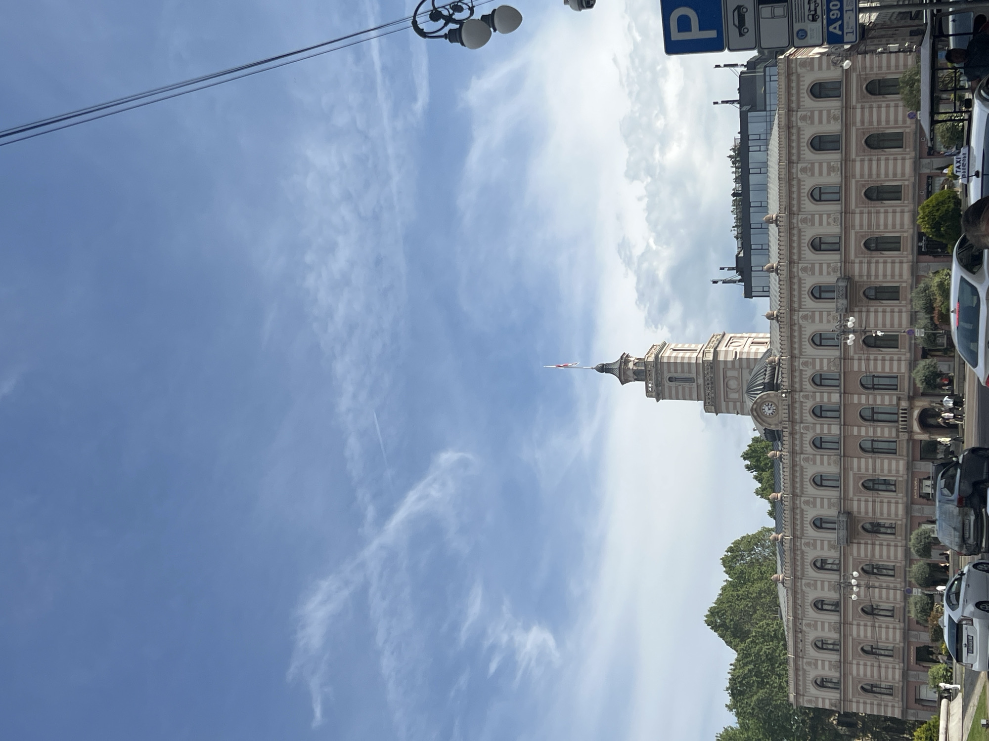 The image shows the clock tower of the Tbilisi City Assembly in Tbilisi, Georgia. The building features a classical architectural style with arched windows, a clock face, and a spire. Trees are located at the bottom edge of the image. Modern glass panels appear to be added to the top of the building. Street lamps and utility wires are visible against a backdrop of a blue sky with wispy clouds. Vehicles are parked along the right side of the frame. Traffic and parking signs are also visible on the right.