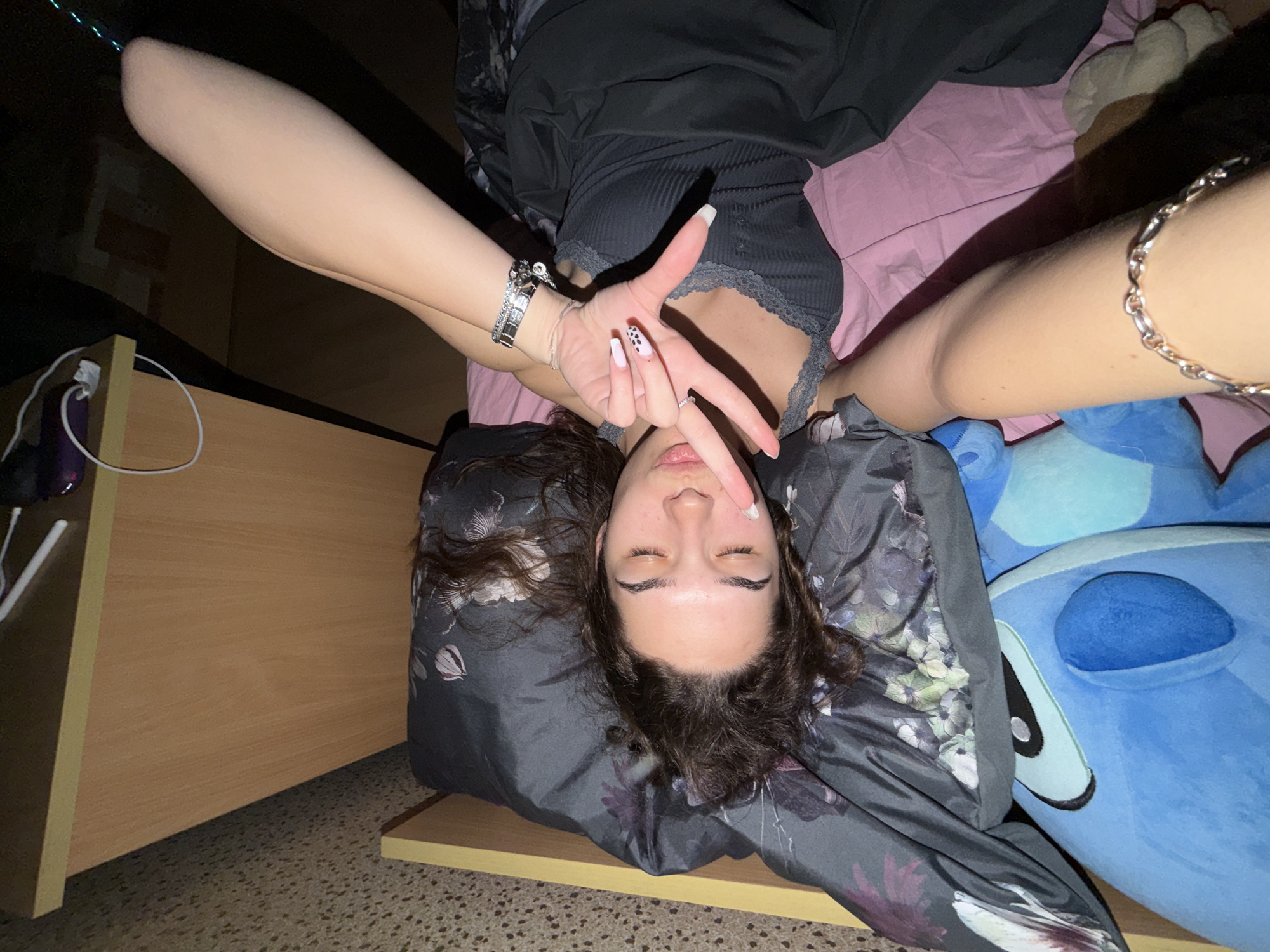 A young person with dark, wavy hair is captured in a close-up, high-angle shot, lying on their back with eyes closed. They are wearing a dark top with lace trim and dark bottoms, partially covered by a duvet with a floral pattern in shades of grey, green, and purple. The person's arms are raised and bent, with the left hand making a V-sign gesture over their face. This hand displays light pink nails, one with black polka dots, and is adorned with multiple silver bracelets and a watch. A silver chain anklet is visible on their right ankle.
The setting appears to be a bedroom, characterized by a pink sheet underneath and a large blue plush toy, resembling the character Stitch, positioned to the person's right. On the left side of the frame, a wooden desk or furniture piece is visible, holding a purple object and a white cable. The lighting is dim, suggesting it is evening or night. The overall mood is relaxed and possibly playful due to the pose. This scene is set in Sofia, Bulgaria. No visible text is present in the image.