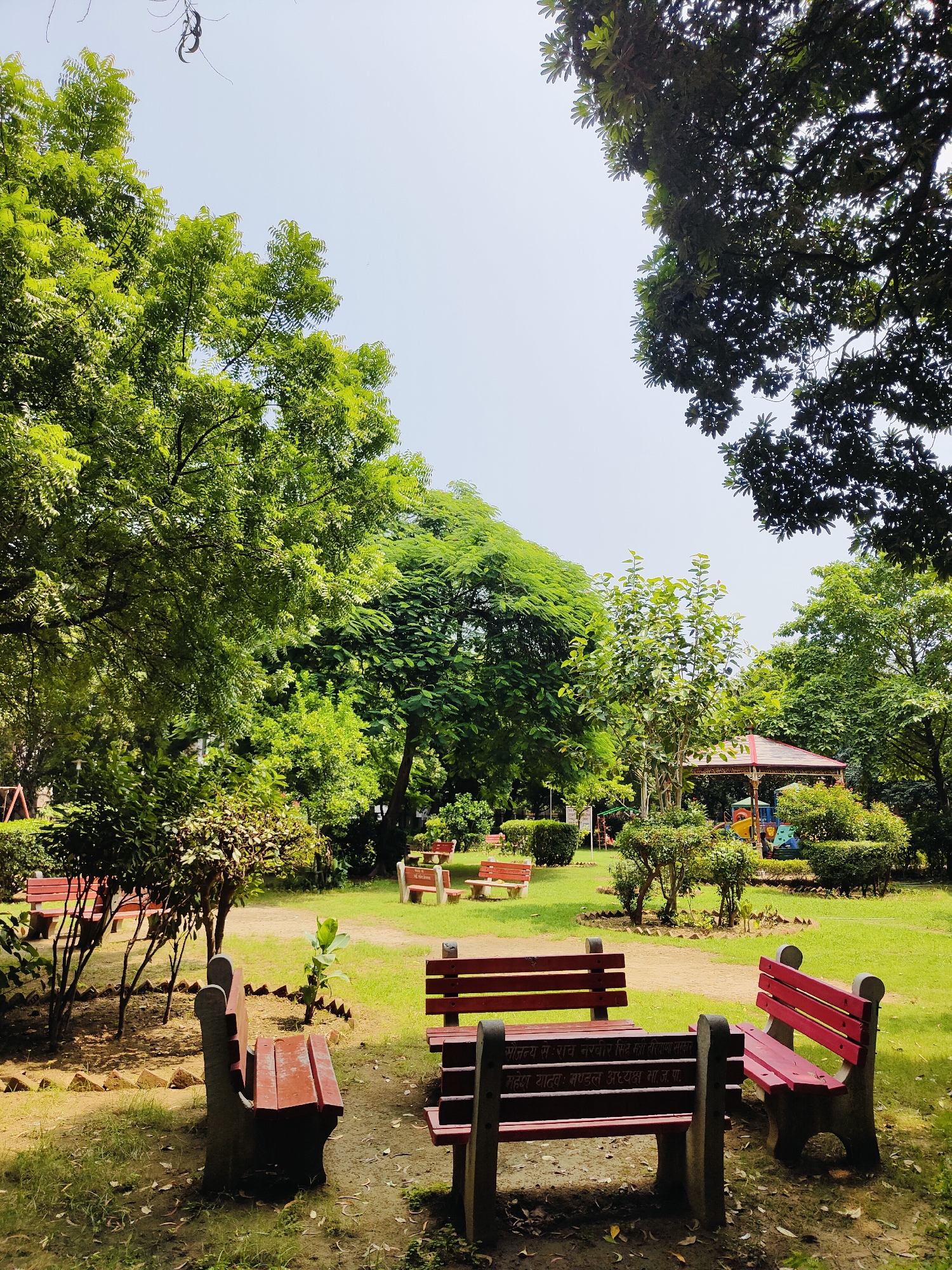 This is a daytime, eye-level, medium shot of a park in Gurgaon, India, likely on a sunny day with a clear sky. In the foreground, there are several red and gray concrete park benches. The bench in the center-right is facing towards the viewer, with its backrest visible. To its left, another bench is positioned at an angle, also with its backrest facing the viewer. In the foreground-left, a third bench is angled towards the left side of the frame, and only its seat is fully visible.
Behind the benches, the park features a grassy lawn with scattered fallen leaves. Several trees with lush green foliage are present, with a particularly large tree dominating the left side of the frame. In the middle ground, a small, octagonal gazebo with a red roof is visible. Around the gazebo, there are more trees and shrubs. To the far left, a playground structure with colorful elements can be seen. The overall impression is of a well-maintained public park in an urban setting.