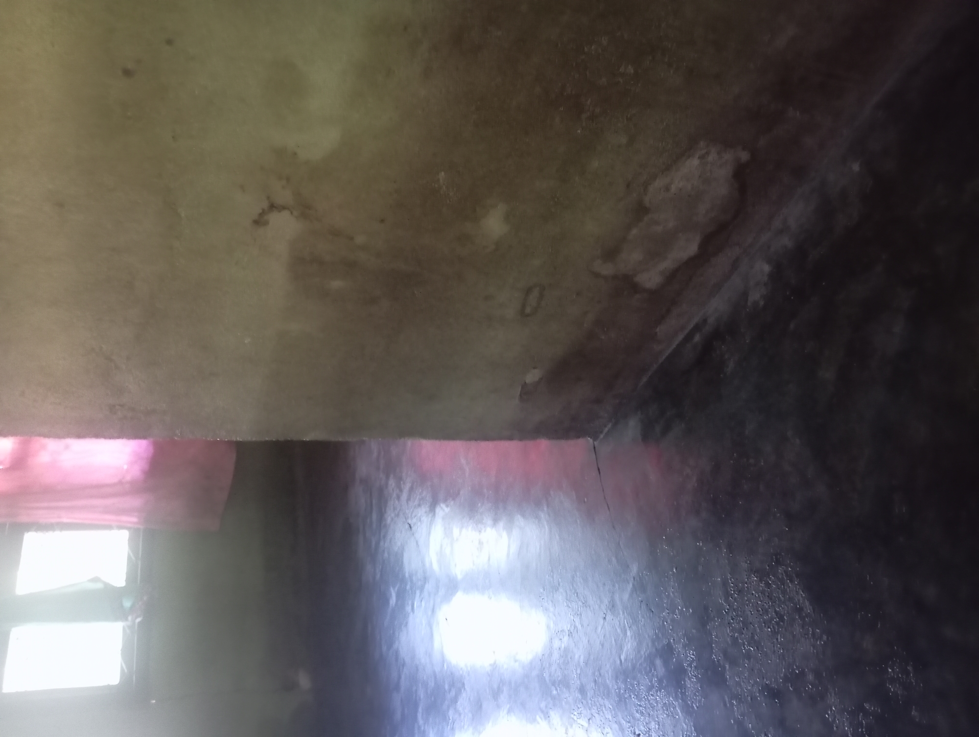 The image shows the interior of a room in Rejosari, Indonesia. The floor is dark and wet, reflecting the light from a window on the left side of the image. A pink curtain hangs near the window. The right side of the image is dominated by a rough, textured wall that appears to be damp and stained. There are patches of peeling plaster on the wall near the floor. The scene suggests an indoor environment that may be experiencing issues with moisture.
