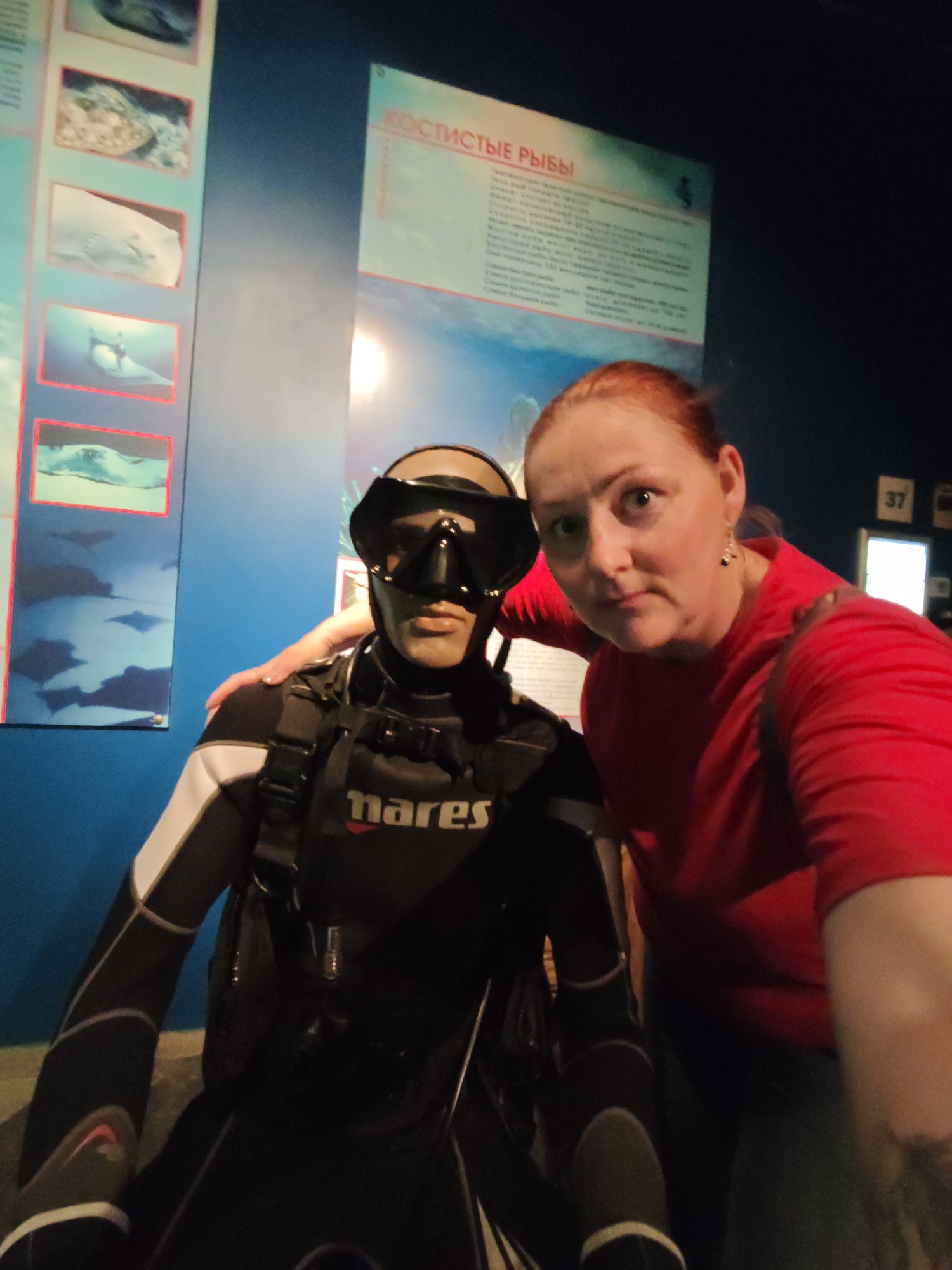A woman with red hair stands with her arm around a mannequin dressed in a black and white wetsuit with a Mares logo. The mannequin wears a black diving mask and appears to be a diver. They are posed in front of a blue wall displaying informational posters with images of marine life. One poster has Russian text at the top. The environment suggests an aquarium or museum setting, possibly the Saint Petersburg Oceanarium. The lighting is dim, typical of such indoor exhibits.