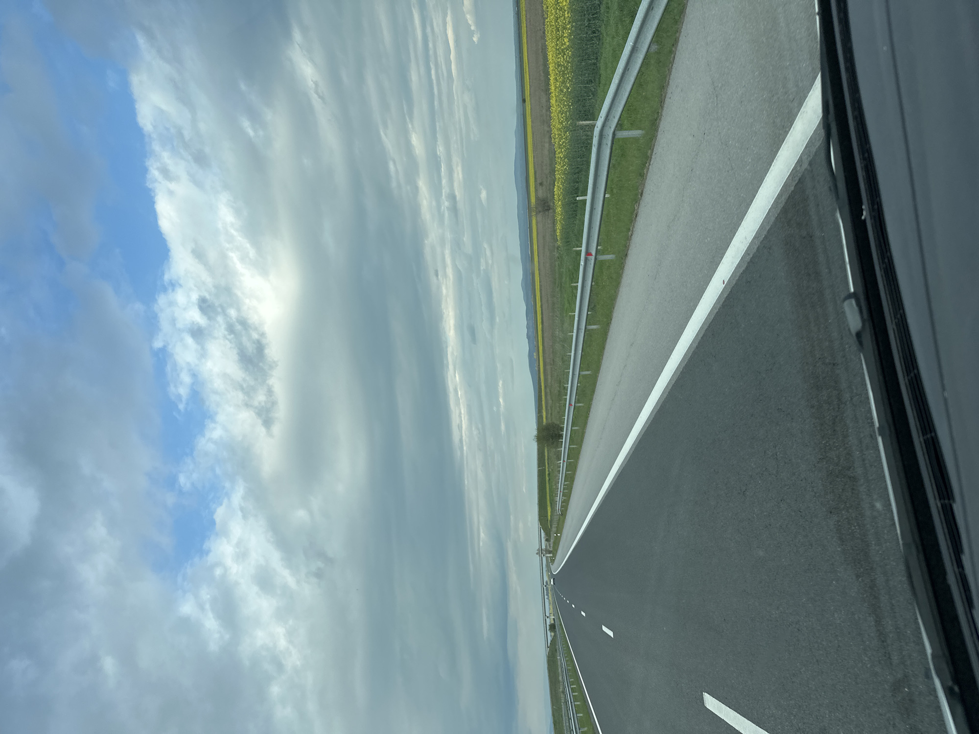 A horizontal view from inside a vehicle of a road in Загорци, Bulgaria shows an asphalt two-lane highway with white line markings. To the right of the road is a metal guardrail and beyond the guardrail is grassy land bordering a field of yellow flowers. The sky is a mix of blue sky and grey clouds. The vehicle's window frame is visible on the right side of the image.