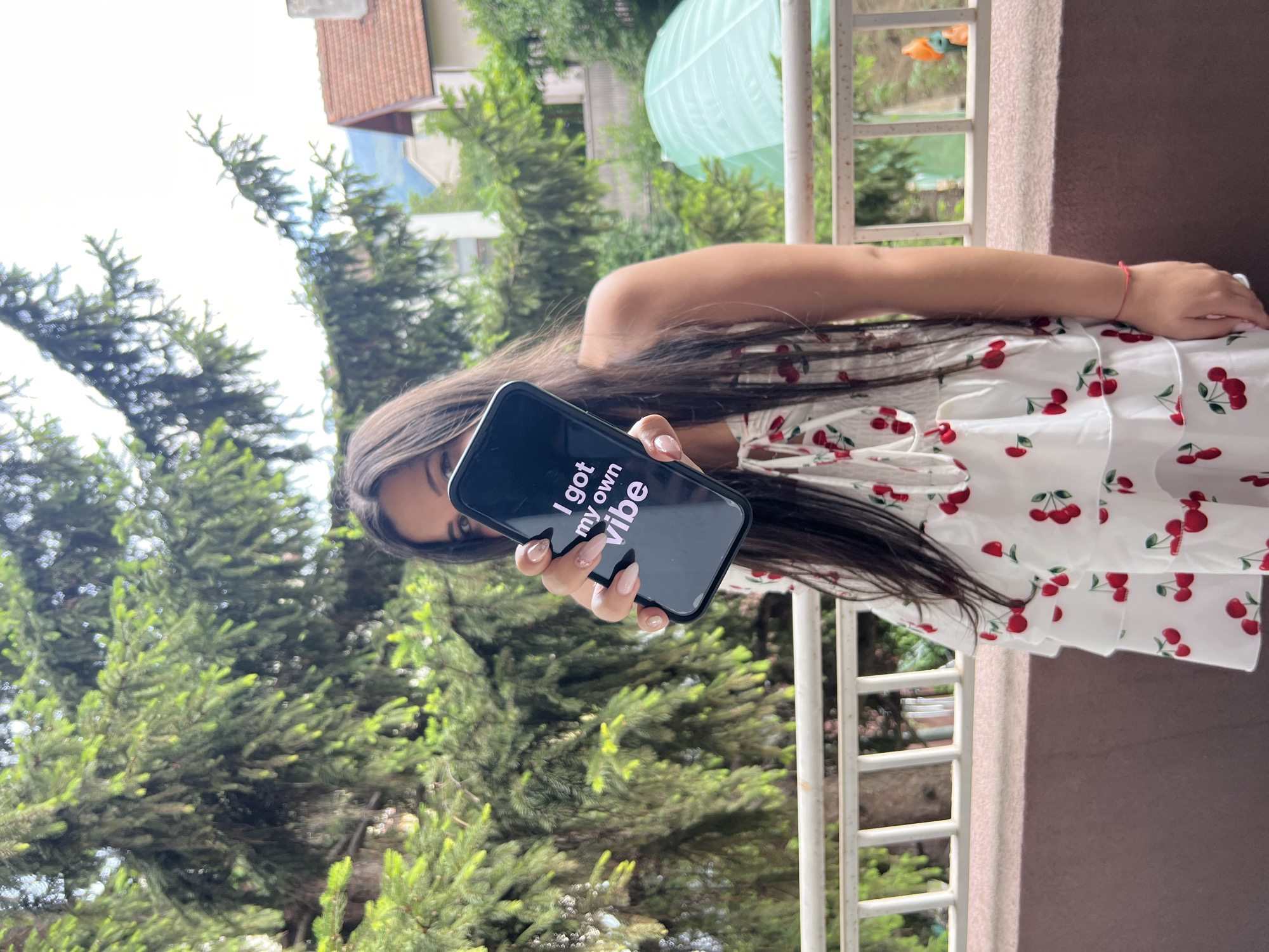 A young woman, whose face is partially obscured by a smartphone she is holding up, stands on a balcony. The phone displays the text "I got my own vibe" in white lettering against a black background. She is wearing a white dress with a pattern of red cherries. Her long, dark hair falls over her shoulders and partially covers the phone. The background suggests an outdoor setting, with lush green trees and the partial view of residential buildings, indicative of a location in the city of Sofia, Bulgaria, likely a residential area with balconies and vegetation. A white railing runs along the balcony. The lighting suggests it is daytime, with bright natural light.
