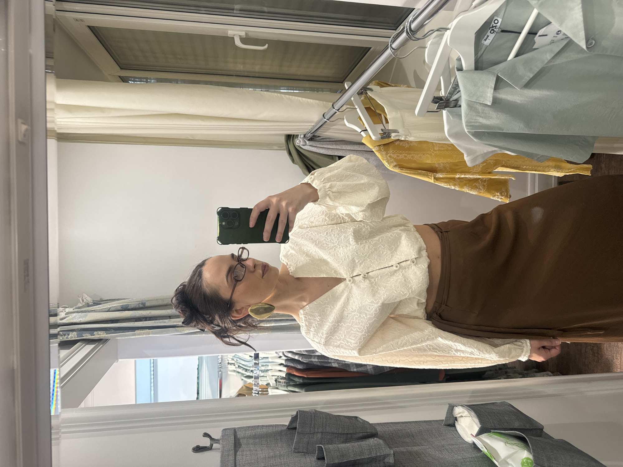 A woman is captured in a mirror selfie, posing in an indoor setting, likely a dressing room or bedroom. She is positioned slightly to the right of the frame, looking towards her left. The woman has dark hair styled back, wears dark-rimmed glasses, and a prominent, flat, golden-toned oval earring on her visible left ear. She is dressed in an off-white or cream-colored blouse featuring puffed sleeves and a textured, possibly embroidered, pattern. The blouse has a row of small, light-colored buttons down the front. Below the blouse, a brown skirt is visible. She holds a dark green smartphone with multiple rear cameras, pointing it towards the mirror to take the photograph.
The background, reflected in the mirror, shows a white window frame at the top with a light-colored blind drawn. Below the window, a silver clothing rack holds various garments on white hangers, including a yellow embroidered top, a white shirt, and a light blue/mint green shirt. A tag on one of the light blue shirts clearly shows the brand name "OKO." To the left, part of a patterned curtain with blue and white designs is visible. At the bottom of the frame, a grey fabric hanging organizer with pockets is seen, one of which contains a package of wipes, with the word "wipes" discernible on its label. The scene suggests a tidy indoor environment.
