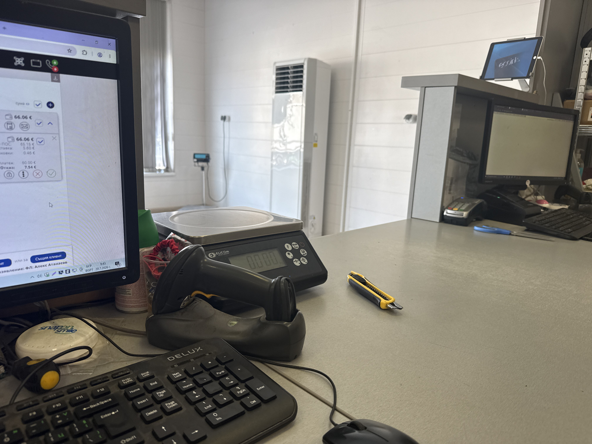 The image shows a checkout counter in a retail store, likely a pharmacy or grocery store, in Rakovski, Bulgaria. The counter is cluttered with various items including a computer monitor displaying a checkout interface, a barcode scanner, a digital scale, a keyboard, a mouse, a credit card reader, and a utility knife. In the background, an air conditioning unit is visible on the wall, along with a tablet device mounted on a stand, displaying the "ecoin" logo. The overall impression is of a functional, operational retail space.