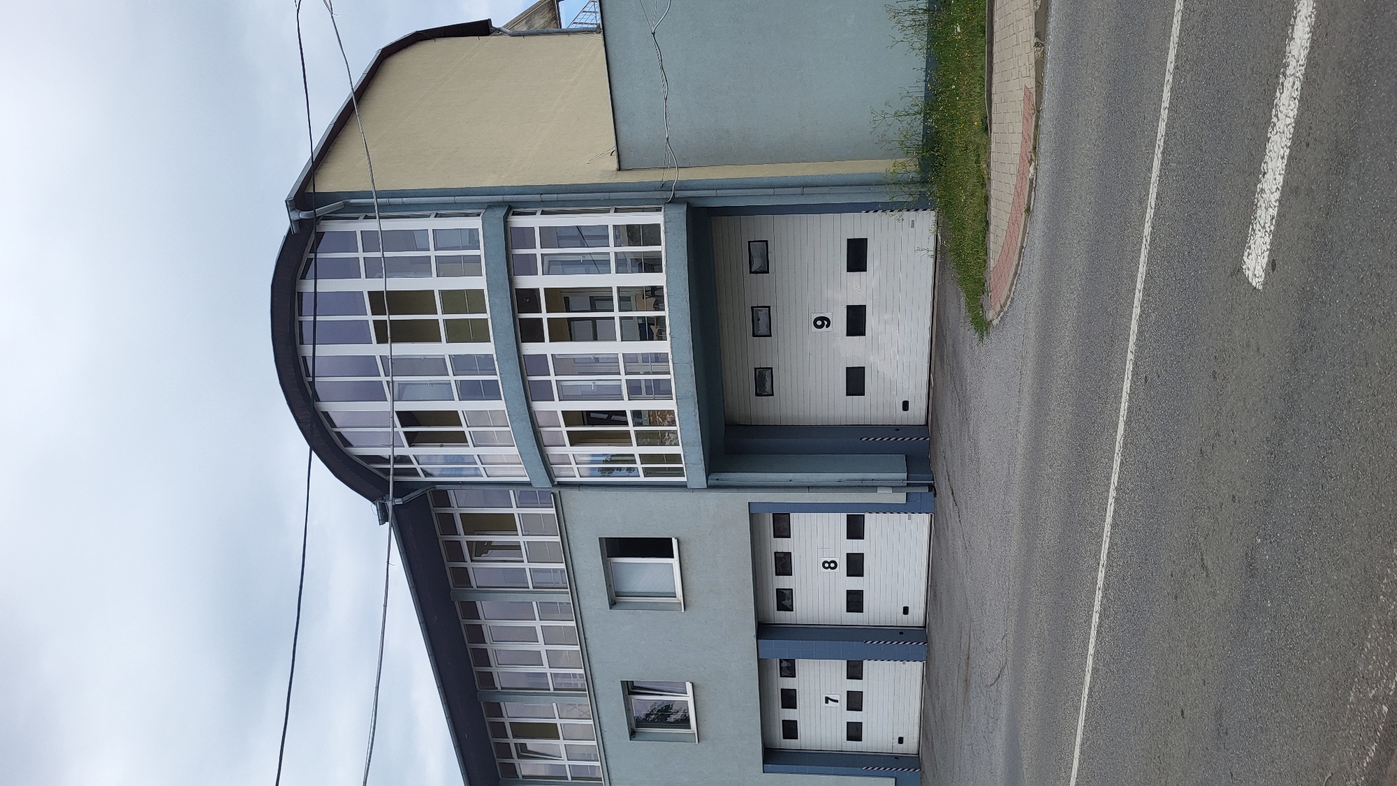 A low-angle, full shot captures a modern, multi-story building with a light blue facade and a beige upper section. The building features a curved, glass-enclosed upper floor that protrudes from the main structure. Below, on the ground floor, are three large white garage doors, labeled with the numbers 7, 8, and 9. The garage doors are partially open, revealing dark interiors. The building is situated next to a grey asphalt road, with a small patch of grass and a brick border visible on the right side. The sky above is overcast with grey clouds. This building is located in the city of Prešov, Slovakia.