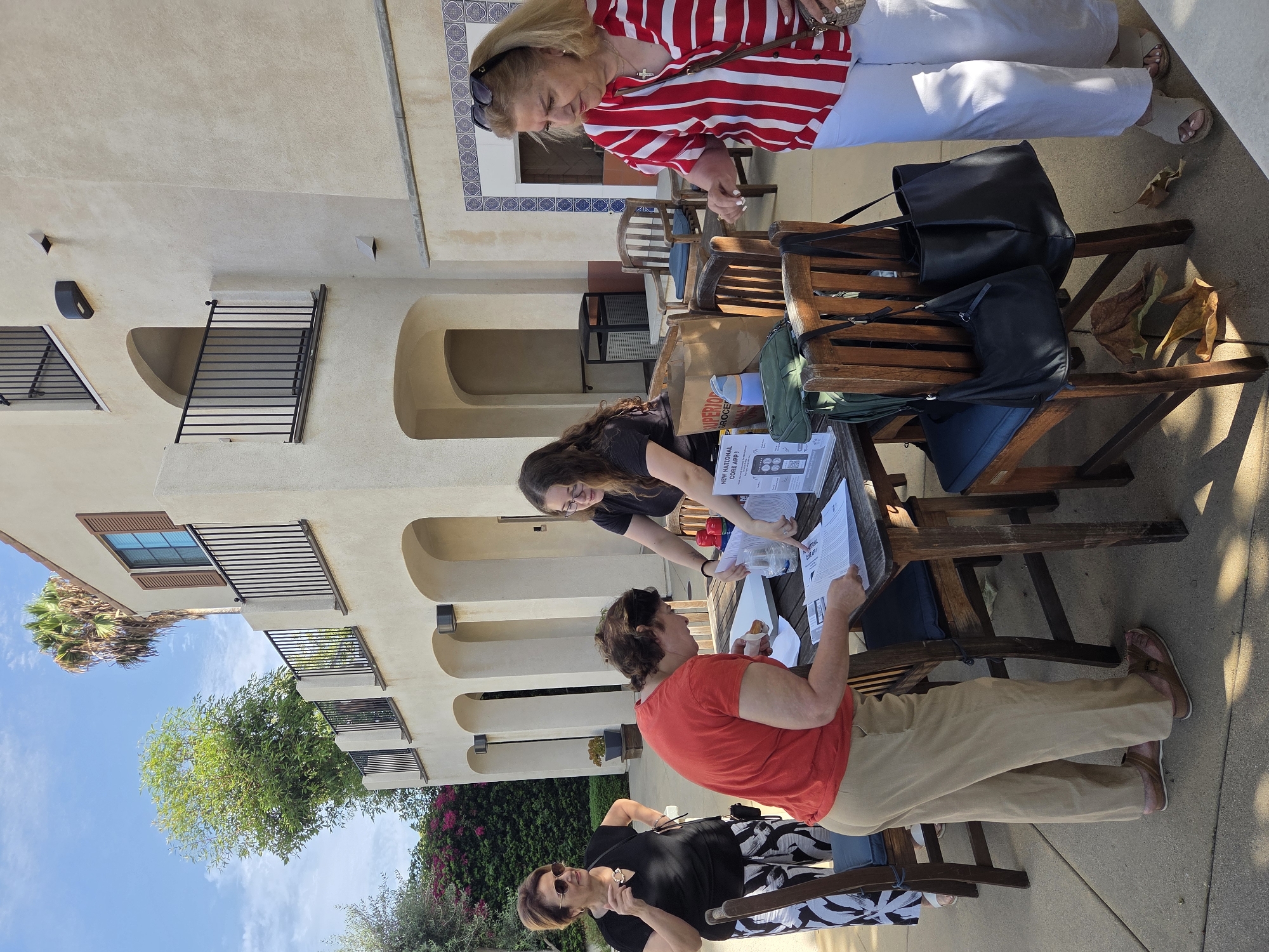 Four individuals are gathered around a wooden table outdoors, situated on a paved surface adjacent to a cream-colored building with arched entrances and balconies. The building has a Mediterranean architectural style, featuring decorative tile work above an arched opening to the right. Green foliage and a palm tree are visible in the background, suggesting a warm climate. The sky is partly cloudy and blue.
On the left, a woman wearing a black t-shirt and black and white patterned pants, with sunglasses perched on her head, stands with her hands on her hips, looking towards the table. She wears a pendant necklace.
At the table, a woman in an orange t-shirt and khaki pants is bent over, looking at papers and holding a small food item in her right hand. She appears to be signing or reviewing documents.
A younger woman with long curly hair, wearing a black t-shirt, stands behind the table, interacting with the woman in orange. She is looking down at the papers and appears to be assisting or explaining something. She is wearing glasses and has a watch on her left wrist.
To the right, a woman in a red and white striped shirt and white pants stands looking at the table. She wears a cross necklace and has sunglasses on her head. She is holding a brown purse on her right shoulder and appears to be in the process of interacting with the group or observing the activity.
On the table are several papers, a plastic water bottle, two red containers, a brown paper bag, a green bag, and a black tote bag. Two dark wooden chairs with dark cushions are placed at the table, with two additional black bags hanging from the back of one chair. A fallen leaf is on the ground near the chairs.
The scene suggests an outdoor gathering, possibly an event, registration, or information session. The location is the city of Glendale, United States of America.