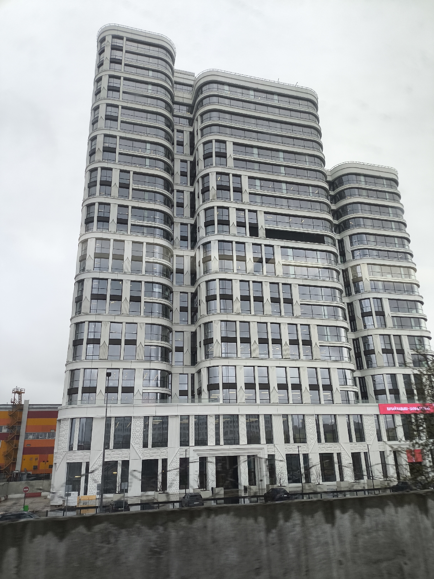 A tall, modern building with a distinctive curved and segmented facade dominates the center of the frame under an overcast, grey sky. The building features multiple interconnected towers of varying heights, all sharing a consistent architectural style. Its exterior is composed of light-colored panels, possibly concrete or metal, interspersed with numerous dark-tinted, rectangular windows that reflect the muted sky. Many window sections are set back from the primary facade, adding depth. The lower levels of the building exhibit more intricate geometric patterns on the light-colored panels, particularly around the entrance area.
In the immediate foreground, a rough, dark grey concrete wall or barrier runs horizontally, obscuring the lowest part of the scene. Behind this barrier, the ground appears wet, suggesting recent rain. Several cars, dark in color, are visible on the wet pavement.
To the left of the main building, another structure with bright orange and yellow panels can be seen, resembling an industrial or commercial building, with a construction crane partially visible near its roof. On the right side of the main building, near its base, a red banner with white Cyrillic text is visible, partially obscured by tree branches. The text appears to read "КАЧЕСТВЕННЫЙ ЗАПА", with the rest cut off. The overall scene depicts a modern urban environment on a cloudy day, emphasizing the building's striking architecture.