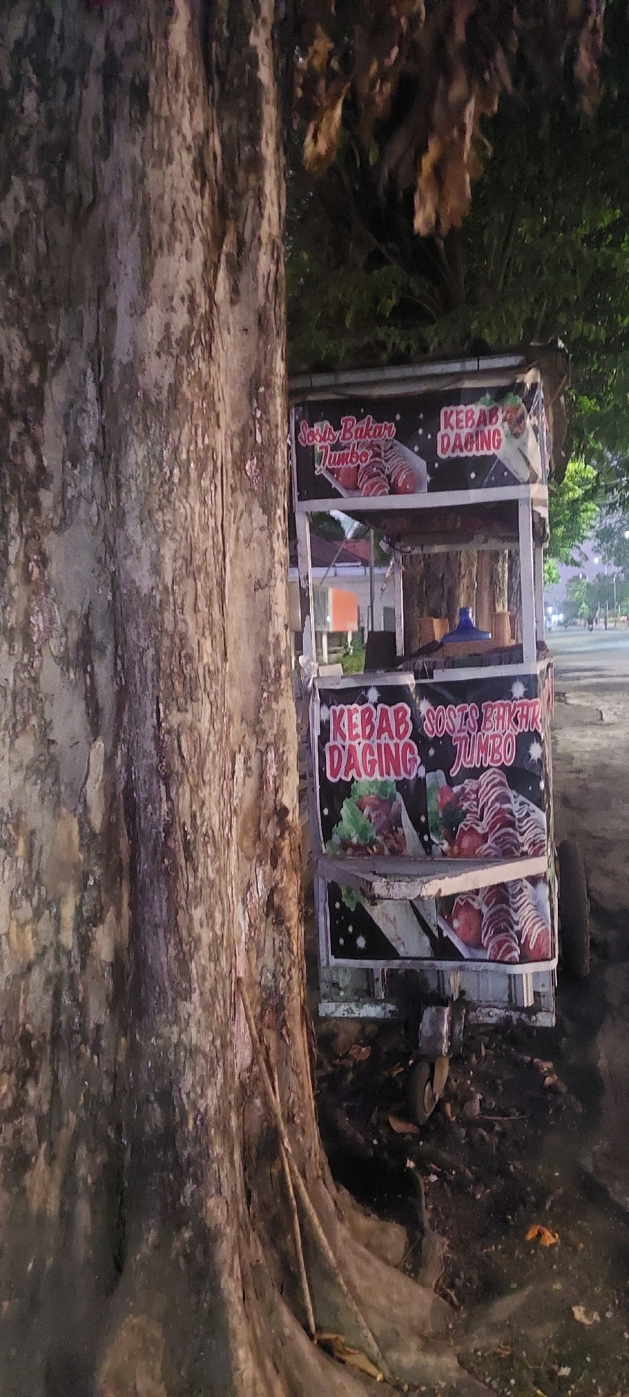 This is a vertical, medium-shot, low-angle photograph taken outdoors at night, likely in the city of Lumajang, Indonesia, as indicated by the presence of a food cart selling Indonesian street food. The foreground is dominated by the textured bark of a large tree trunk on the left side of the frame. To the right of the tree is a mobile food cart, which appears to be for selling "Kebab Daging" (meat kebab) and "Sosis Bakar Jumbo" (jumbo grilled sausage). The cart has a white base with black signage displaying images of the food. Above the cart, there is a roof structure made of dark fabric. The background shows a dimly lit, paved area, possibly a street or parking lot, with some distant trees and streetlights visible. The overall impression is that of a common street scene at night in Indonesia, with a focus on the food cart as a subject.