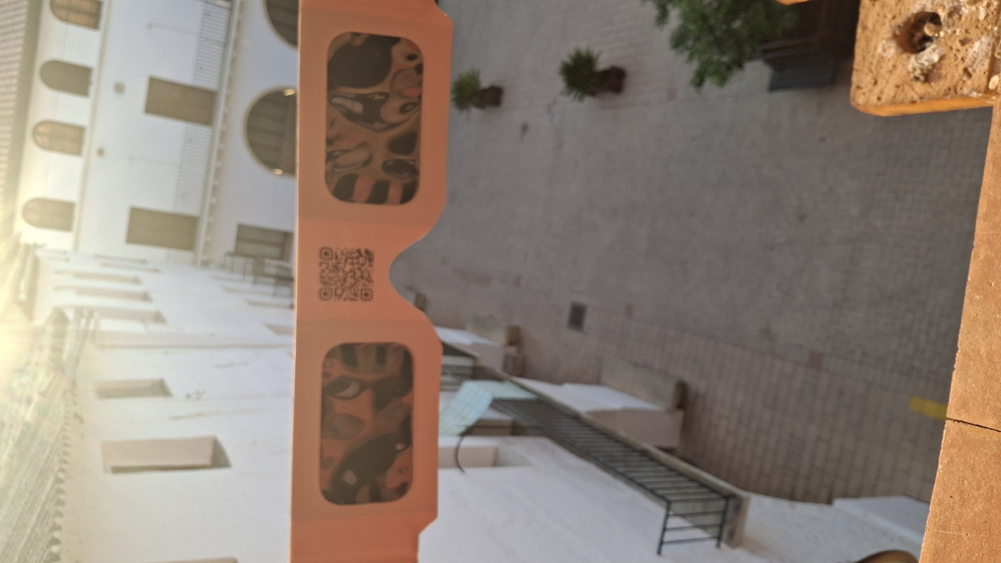 A pair of orange eclipse glasses are held up in the foreground, obscuring a view of a white building with arched windows and doorways and a tiled roof. The glasses have a QR code printed on the left side and show distorted images of a solar eclipse through the lenses. In the background, a courtyard with a brick-paved floor is visible, with a black metal railing on the left and two potted plants. The scene appears to be set in the town of Híjar, Spain, known for its historically significant architecture and astronomical events.