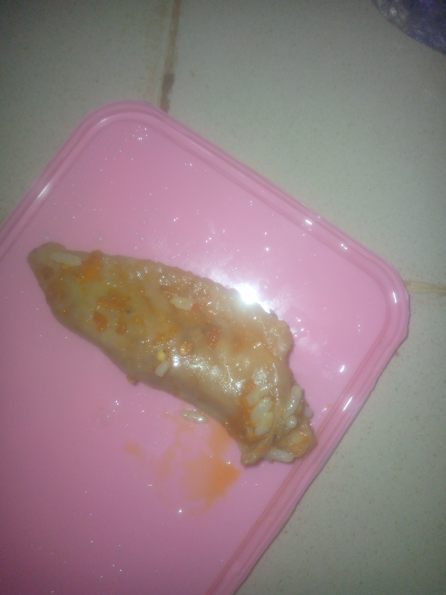 The image displays a single, elongated piece of food resting on a pink plastic tray or container lid. The food item is light brown or beige, coated in a glossy, somewhat thick sauce with visible orange-red specks and white rice grains embedded within or sticking to its surface. It resembles a stuffed vegetable, such as a dolma, or possibly a piece of meat like a chicken wing prepared with a saucy coating.
The pink container is rectangular with rounded corners and a raised edge, suggesting it's designed to hold food. There are shiny reflections on its surface, possibly from moisture or oil from the food. The container is placed on a light-colored, possibly tiled, surface that appears to be a floor or counter. Visible lines on this background suggest grout or seams between tiles.
The lighting is somewhat dim, and the image quality is soft and slightly grainy, hinting at an indoor setting, likely taken with a mobile phone camera in moderate light. No activity is explicitly depicted, but the arrangement implies the food is prepared for consumption or serving. There is no visible text.