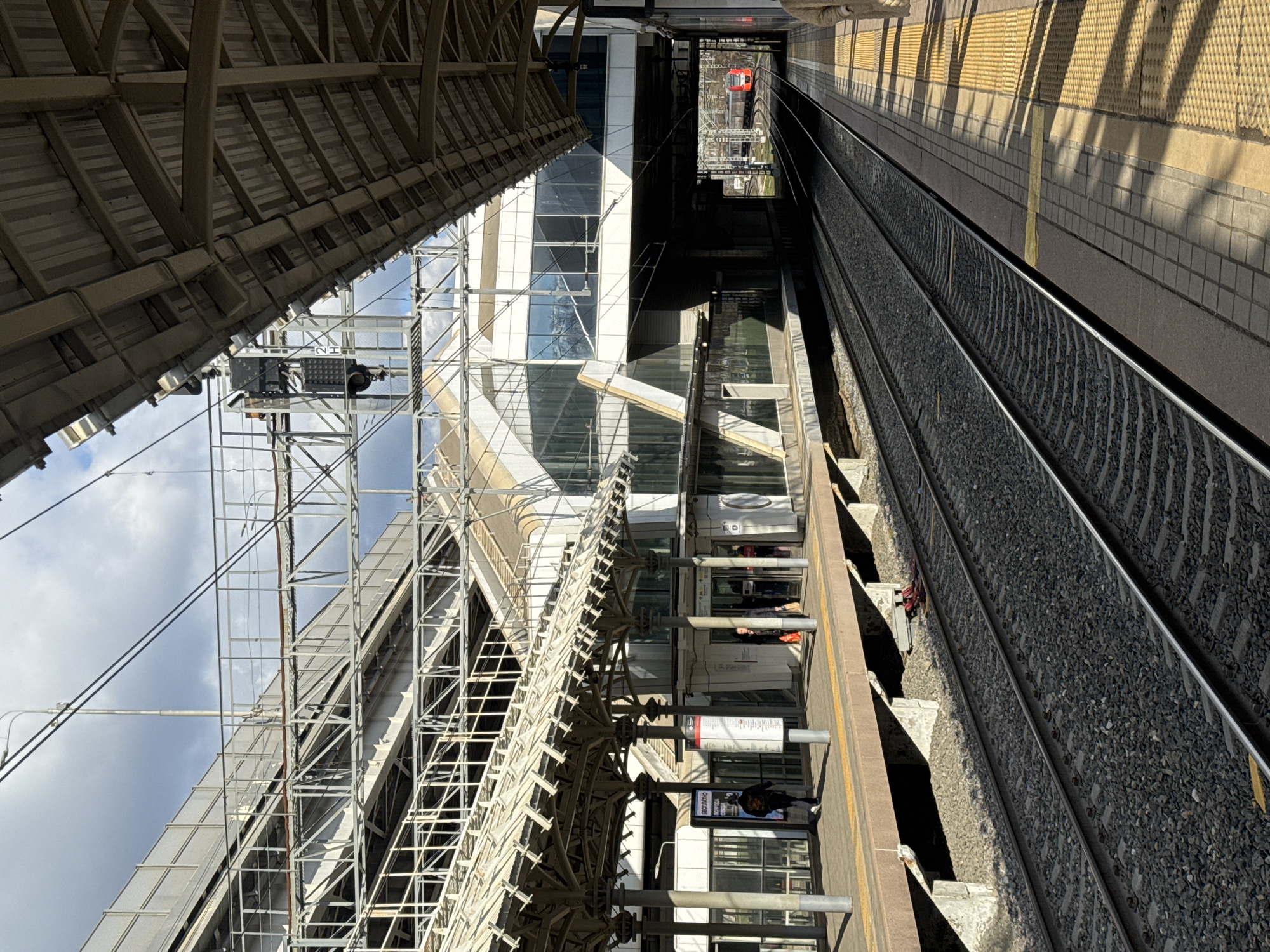 The image depicts an elevated view of a train platform and tracks in Moscow, Russia. The platform runs along the left side of the frame. Structures and supports, some under construction, tower above the platform, including scaffolding, a metal roof, and a building with glass panels. People are visible on the platform, near signage and in the building behind the platform. Train tracks occupy the right side of the frame, with the platform edge running parallel. Across the tracks, there is a yellow textured section of the platform. A maintenance crew can be seen in the distance working on the rail.