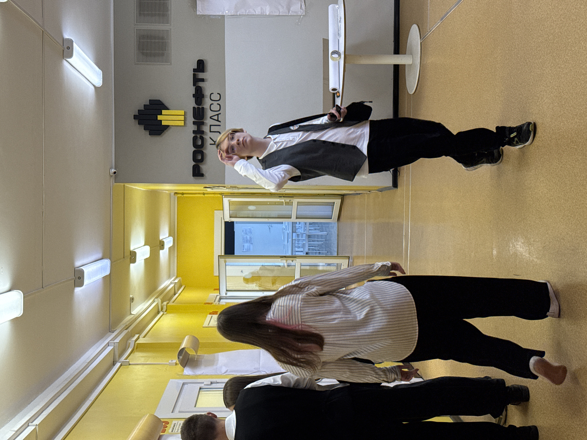 A hallway in a building in Krasnoyarsk, Russia, is shown. On the right side, a student with blonde hair, glasses, and wearing a white shirt with a black vest and black pants stands next to a round table holding a roll of paper. The wall behind the student is grey and features the words "Роснефть КЛАСС" in Russian. Another student wearing a striped shirt and black pants walks towards an open doorway in the middle of the hallway. Another student is visible between the first two. The hallway has yellow walls, and fluorescent lights are affixed to the wall on the left. The floor is yellow. There are tables along the hallway.