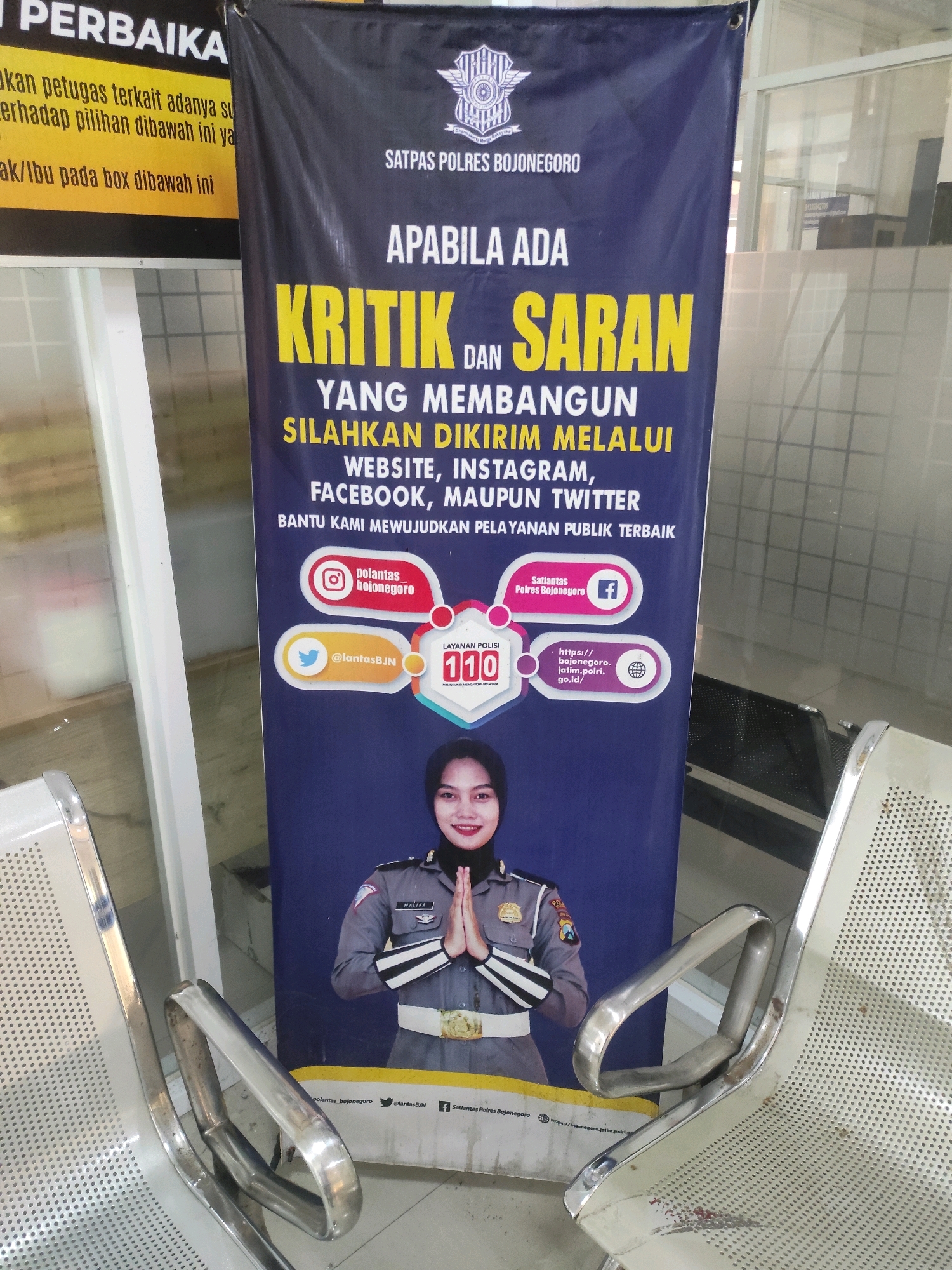 The image is a close-up shot of a banner displayed vertically on a dark blue background. The banner, placed in what appears to be a waiting area with metal chairs, contains text and logos in yellow and white. At the top of the banner, text in white reads "SATPAS POLRES BOJONEGORO." Below this, in large yellow letters, it says "APABILA ADA KRITIK DAN SARAN." Further down, in white text, is "YANG MEMBANGUN SILAHKAN DIKIRIM MELALUI WEBSITE, INSTAGRAM, FACEBOOK, MAUPUN TWITTER." The banner also features several social media icons and contact information, including an Instagram icon labeled "polantas_bojonegoro," a Twitter icon labeled "@lantasBJN," a Facebook icon, and a globe icon with a web address. At the bottom of the banner, a woman in a police uniform with a headscarf is depicted with her hands pressed together in a gesture of greeting. The overall context suggests this is an announcement from the Bojonegoro Police Department in Indonesia regarding public feedback channels.