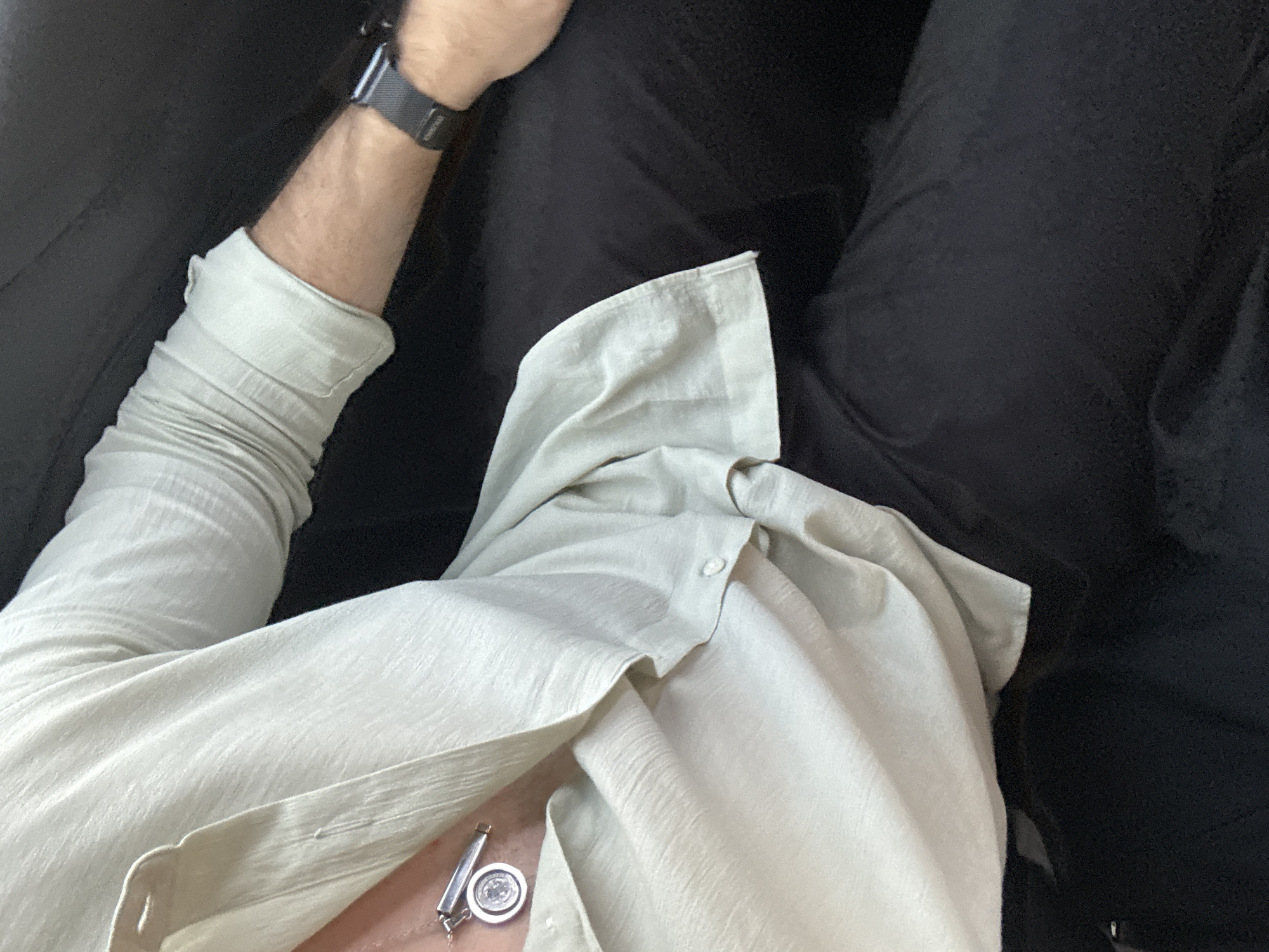 A close-up, overhead shot shows a person seated. The individual is wearing a light-colored, button-down shirt with rolled-up sleeves. On the left wrist is a dark-colored smartwatch. A necklace with a silver pendant is visible. The person is wearing dark-colored pants. The background is dark, possibly upholstery or clothing. The image does not contain enough information to determine a specific location in San Bernardino Tlaxcalancingo, Mexico.