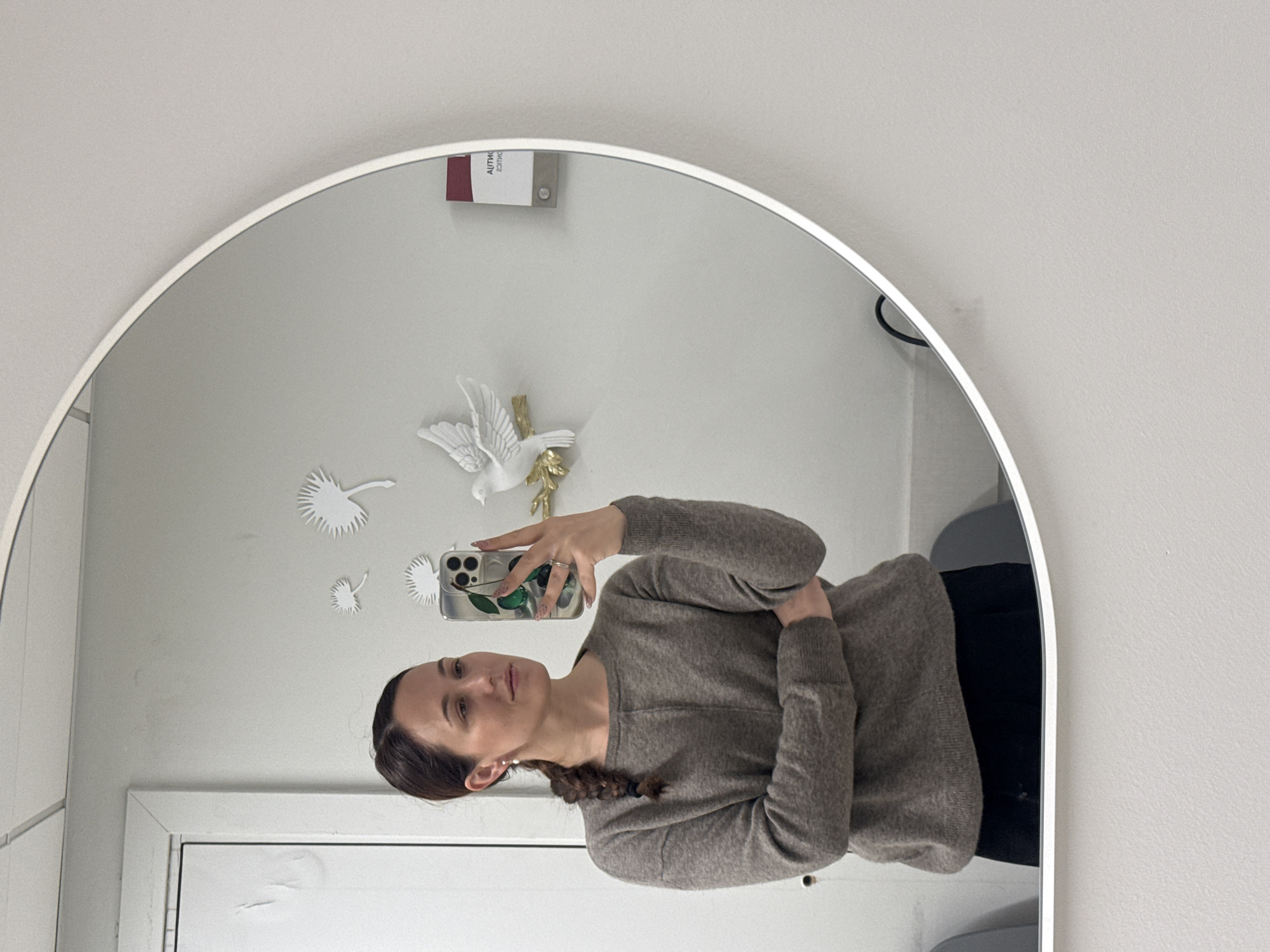 A person stands in front of an oval mirror, taking a selfie with a smartphone. The person is a woman with brown hair styled in a braid and wears a brown sweater and black pants. The mirror reflects a white wall adorned with decorative paper cutouts shaped like leaves or feathers and a small white bird figurine with gold accents. A white door with a window is visible at the bottom of the mirror. The lighting appears to be indoor, and the environment is plain and modern. The location is not discernible from the image alone and could not be determined to be in Riga, Latvia.