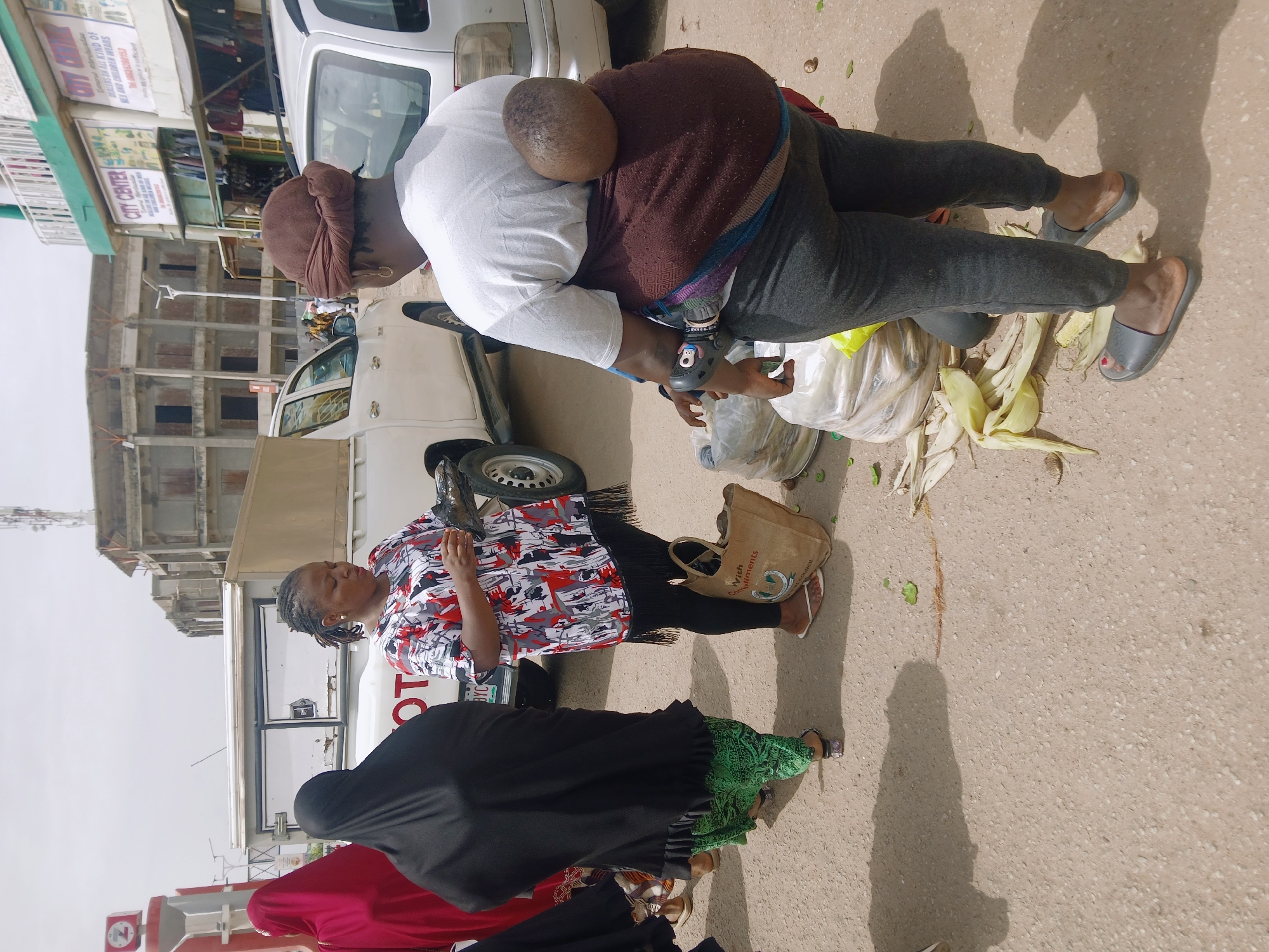 This image shows a street scene in Jos, Nigeria. In the foreground, there are three women and a child in a sling on the back of one of the women. The first woman on the right is wearing a black robe with a green patterned hem. The second woman is wearing a red and white patterned shirt with black leggings. The woman carrying the child is wearing a white long-sleeve shirt with grey leggings and grey slip-on shoes. She also wears a brown head covering. The child is wearing a brown sweater. The woman is carrying a tan tote bag with the word "With" and "Council" on it. There is also what appears to be corn husks and some plastic bags on the ground next to them. There are white trucks parked near the women. In the background, there is a partially completed building with a sign that reads "CITY CENTER" and lists it as a "DEALER ON ALL KIND OF KEY AND CHILDREN WEARS." The ground surface is light brown.