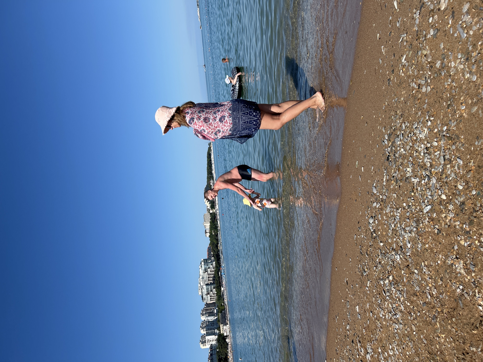 This image shows a beach scene in Gelendzhik, Russia. In the foreground, a person wearing a pink bucket hat and a patterned cover-up stands by the water's edge, facing left. To their left, a man in black swim trunks and a young child in a swimsuit and yellow hat are in the shallow water. The man is holding the child's hand, and they appear to be interacting. The water is clear and calm, with a sandy bottom visible. In the background, there is a shoreline lined with buildings and trees. Other people are visible in the water and on the beach, some on inflatable rings. The sky is clear and blue. The beach is covered in sand and small pebbles.