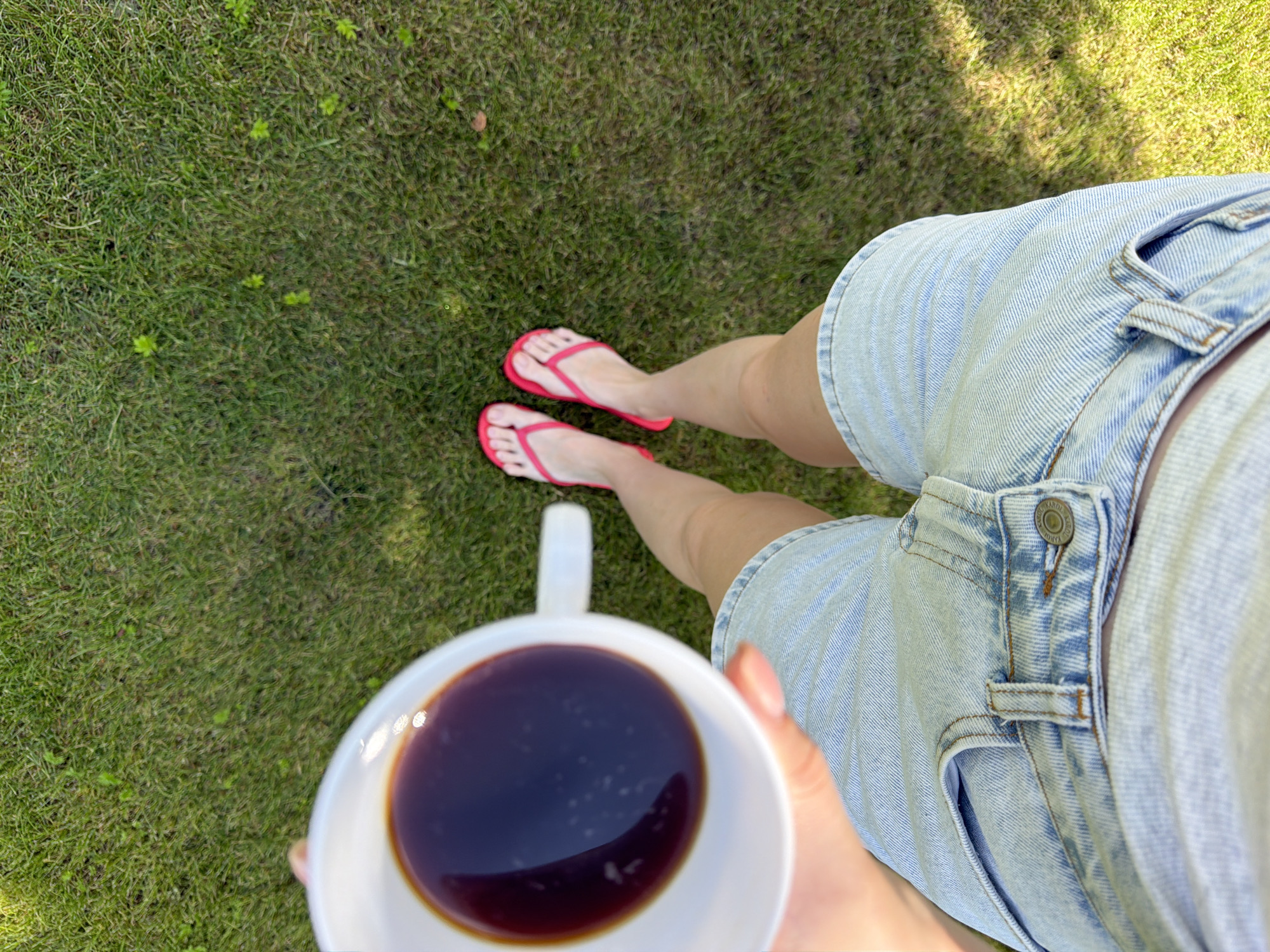 An overhead shot shows a person holding a white mug of dark liquid, presumably coffee, while standing on a patch of green grass. The person is wearing light blue denim shorts and bright pink flip-flops. The camera angle looks down at the person's legs and feet, then up towards the mug. The background consists of a grassy lawn, with some areas in sunlight and others in shadow, suggesting an outdoor setting in a park or backyard. There is no indication of the specific location within Miasskoye, Russia.