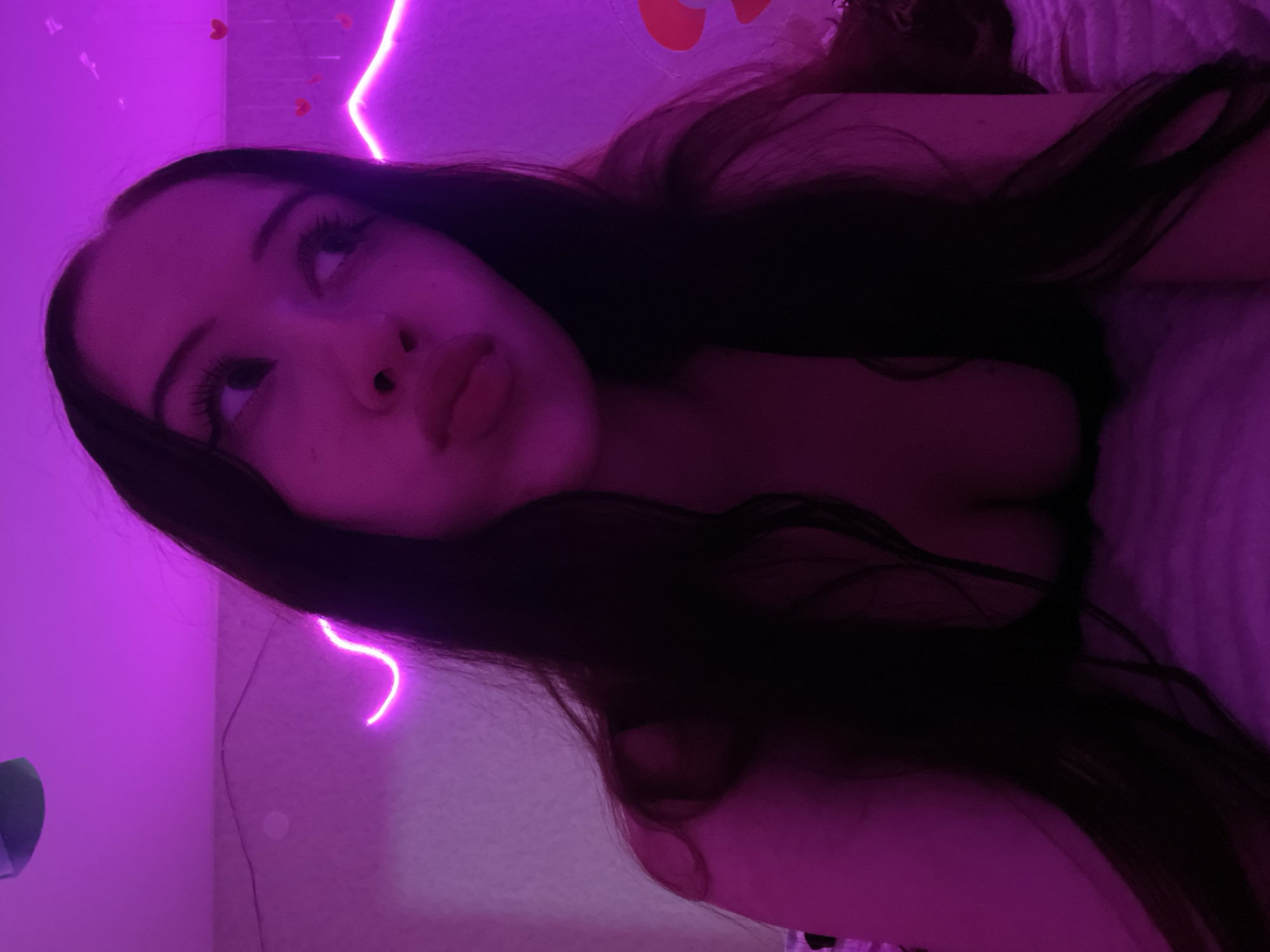 The image is a close-up, low-angle, indoor selfie taken in a room illuminated by purple neon lights. A young woman with long, dark hair is the central subject, positioned on a bed with a textured pink surface. She is looking away from the camera with her lips pursed. The background features a pink wall with neon lights forming a wavy pattern and some small heart decorations. The overall impression is of a dimly lit, intimate personal space, potentially a bedroom.