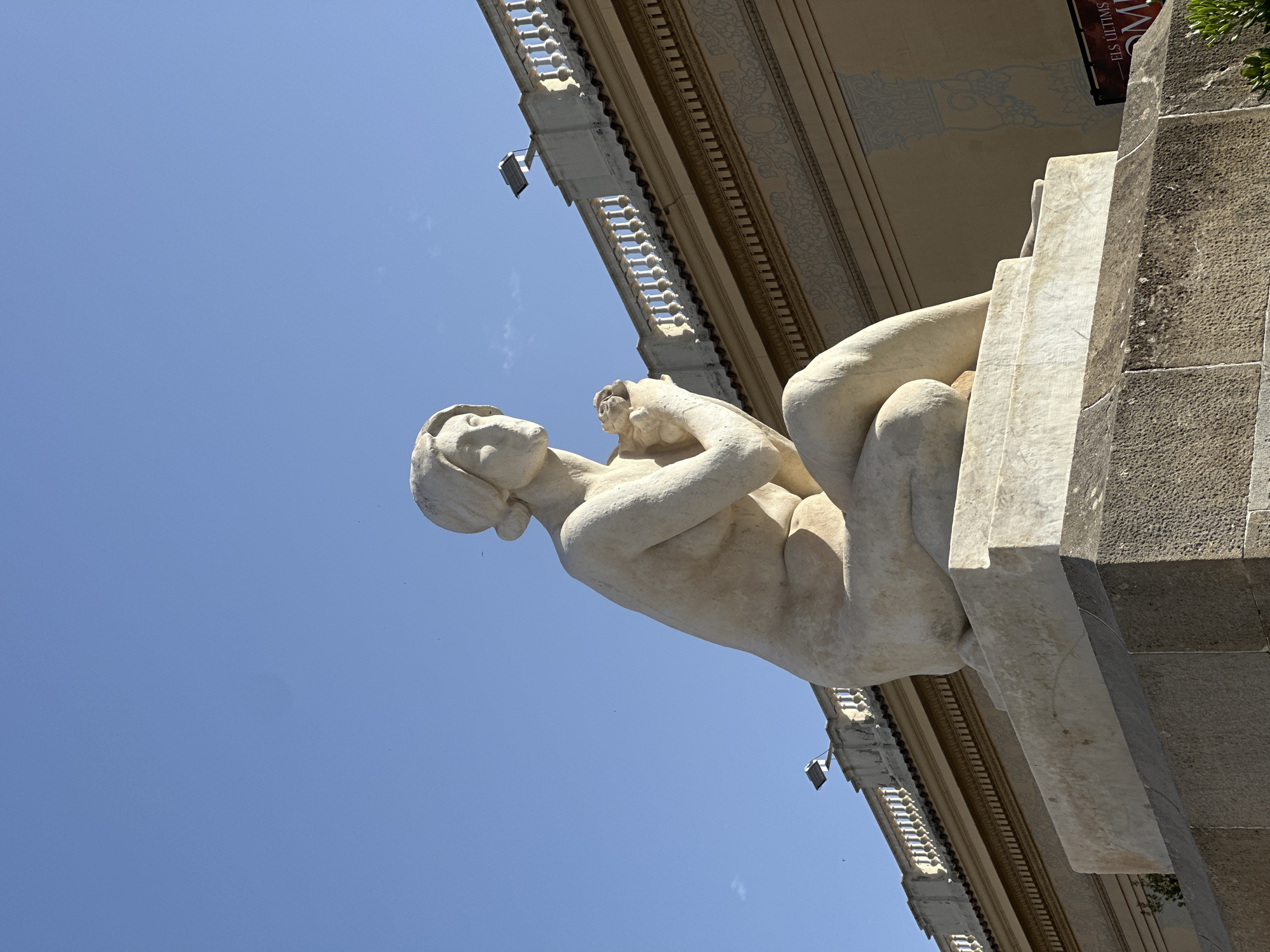 The image shows a life-size statue of a woman carved from marble, seated on a stone pedestal in front of a building. The statue depicts the woman with her knees drawn up, cradling a small object in her hands, and looking off to the side. She has her hair pulled back in a bun. The building behind her is light brown with decorative friezes and a white balustrade along the roofline. Two lights are mounted on the roof. The sky is a clear, bright blue. A sign in the lower right corner reads "ELS ÚLTIMS EM". This statue is located in the Parc de la Ciutadella in Barcelona, Spain, specifically in the Cascada Monumental.