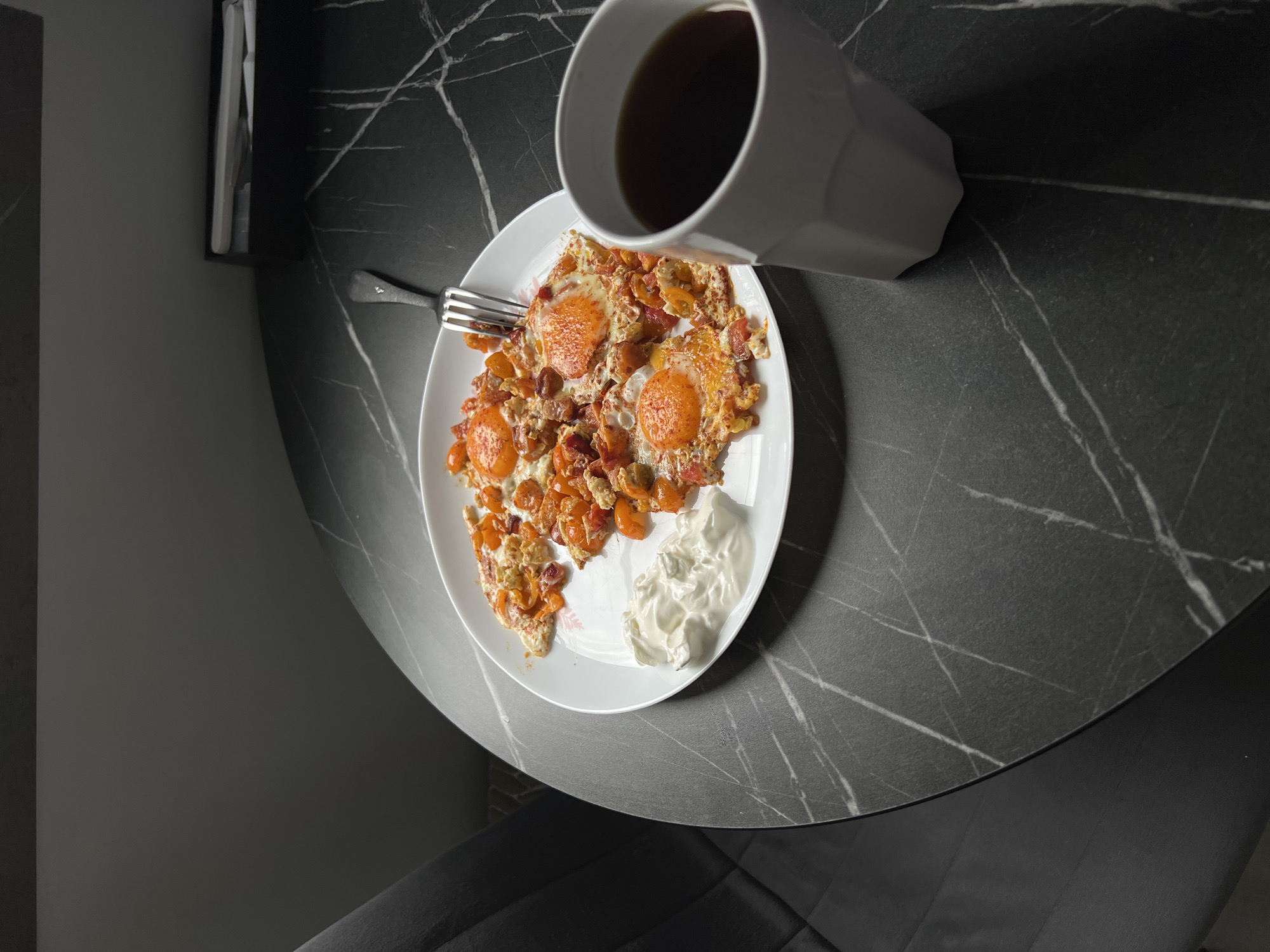 A high-angle, medium shot depicts a partially eaten meal on a white plate on a dark, marbled table. The meal appears to consist of fried eggs with cooked tomatoes and an unidentified white creamy substance, likely sour cream or yogurt. A metal fork rests on the plate near the food. A white, angular mug filled with a dark liquid, likely coffee or tea, is positioned near the plate. The setting appears to be indoors, possibly a dining area, with dark walls and a chair visible in the lower part of the frame. The location is not evident in the photo, so it is impossible to confirm it's in Khimki, Russia.