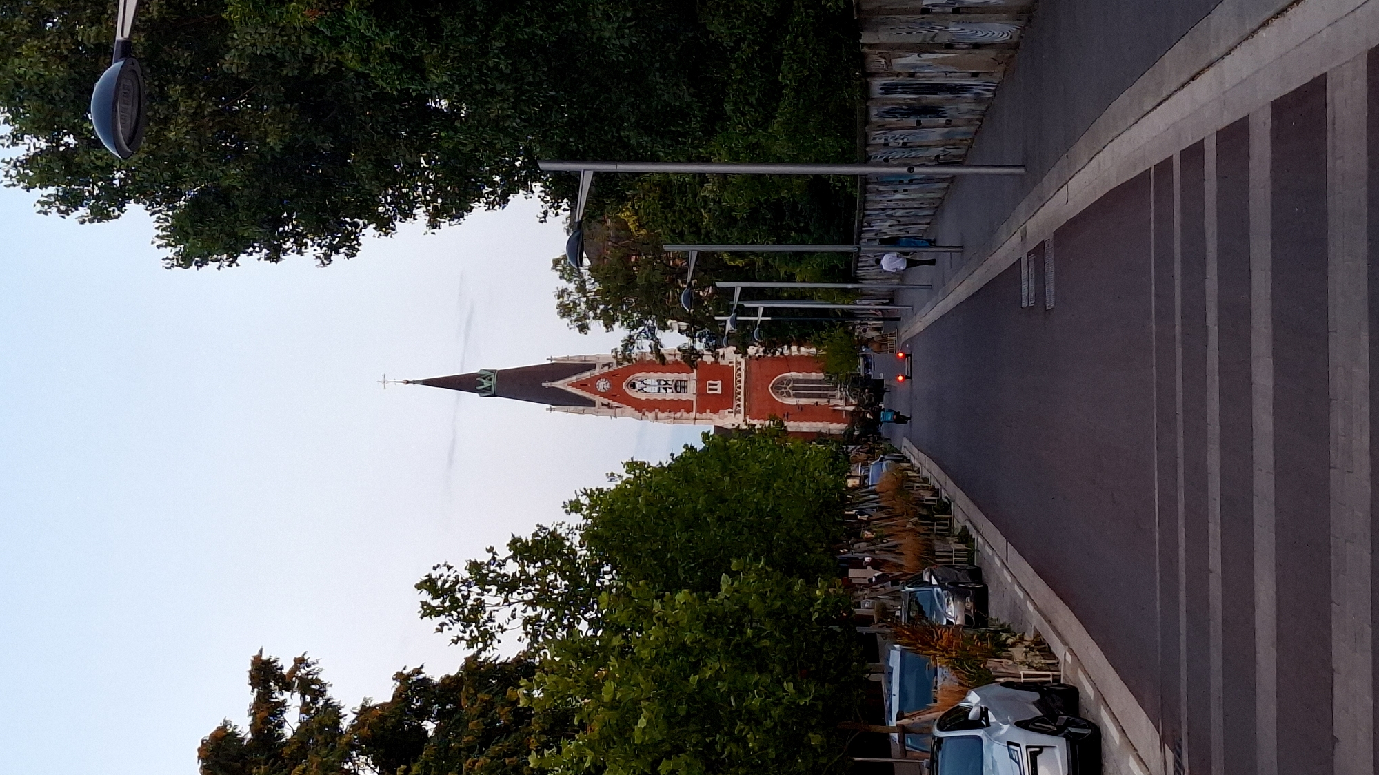 The image shows a street scene in Vienna, Austria, with a prominent red brick church tower with a steeple in the background. The church is flanked by trees on either side of the street, and there are streetlights visible. Cars are parked along the left side of the street, and pedestrians are walking on the right side. A crosswalk is visible in the foreground. The sky is overcast, suggesting it may be dusk or dawn. The overall impression is that of a calm, urban environment with a strong architectural presence.