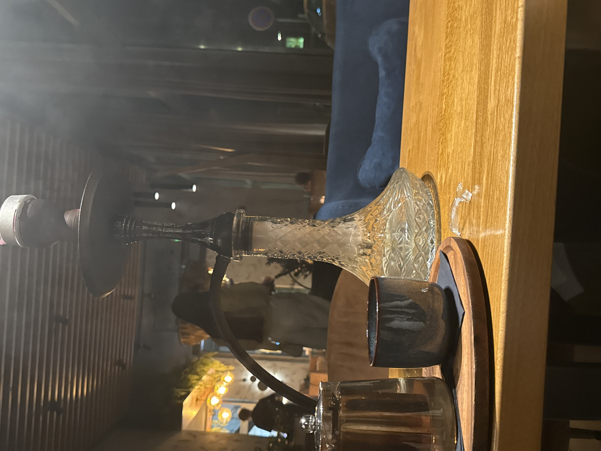 In a dimly lit, modern establishment, likely a shisha bar or lounge, in Bratislava, Slovakia, a hookah stands as the central subject. Smoke curls from its glass base, a testament to recent use. In the foreground, a wooden tray holds a dark ceramic cup and a glass teapot with a metallic lid. The table itself is made of polished wood. In the background, blurred figures move through the space. A blue upholstered bench is visible to the right, and through large windows, the evening city lights and streetlights of Bratislava can be seen, including a circular street sign and a parked vehicle. The overall ambiance is one of relaxed evening leisure.